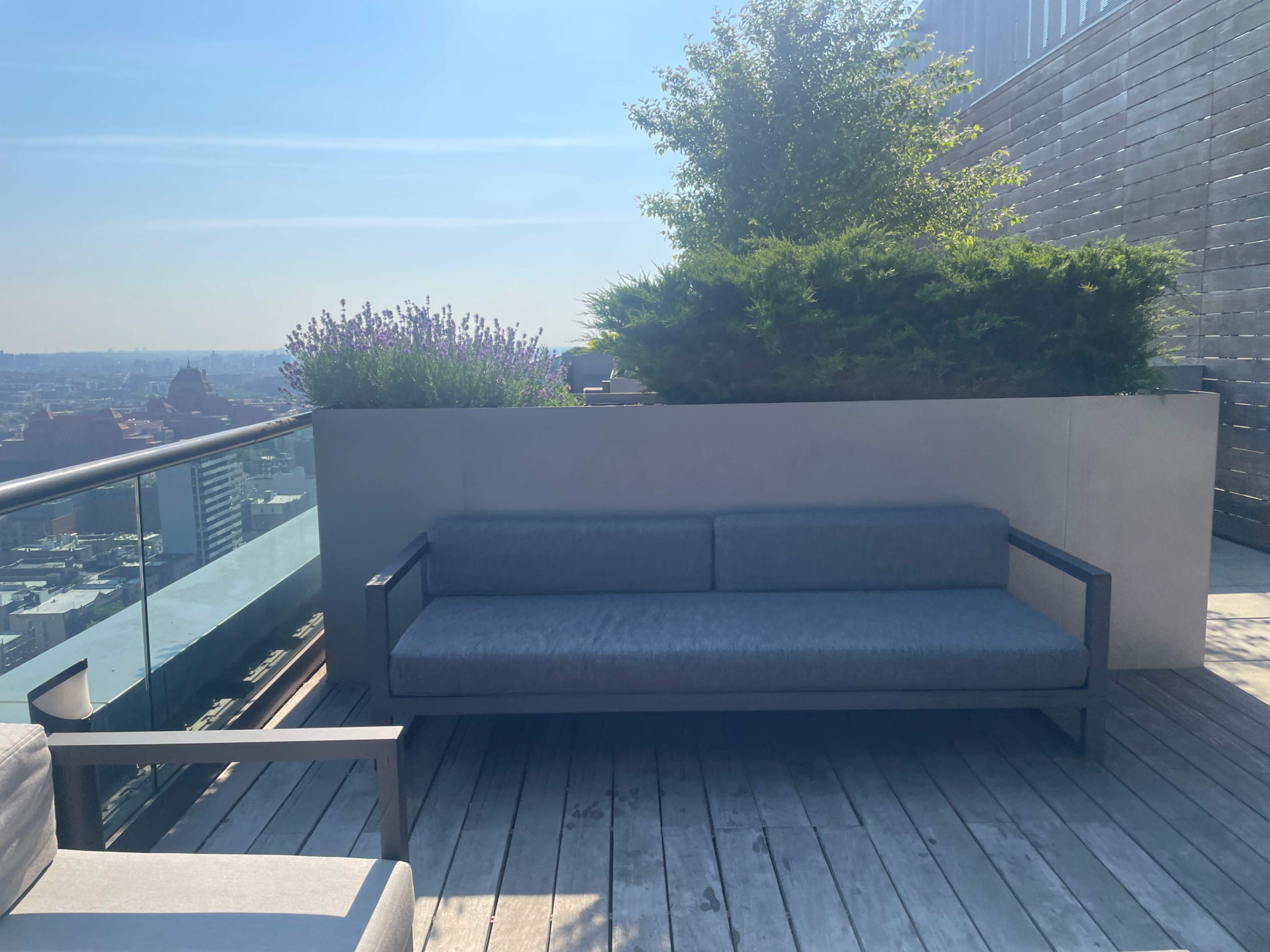 A modern rooftop terrace with a gray sofa, wooden decking, and potted greenery against a clear blue sky.