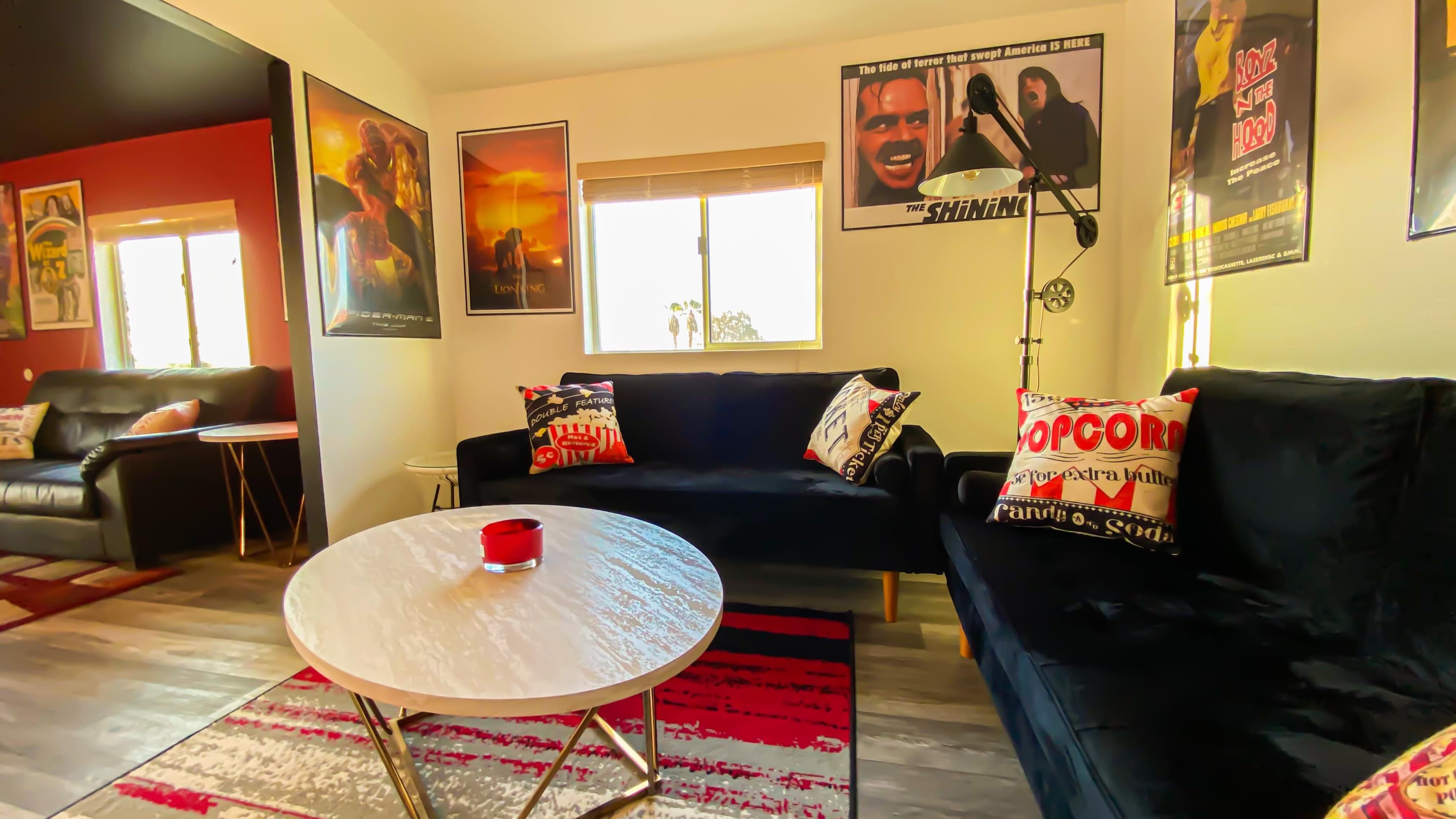 The scene features a living room with two black sofas, a round coffee table, and several movie posters on the walls.