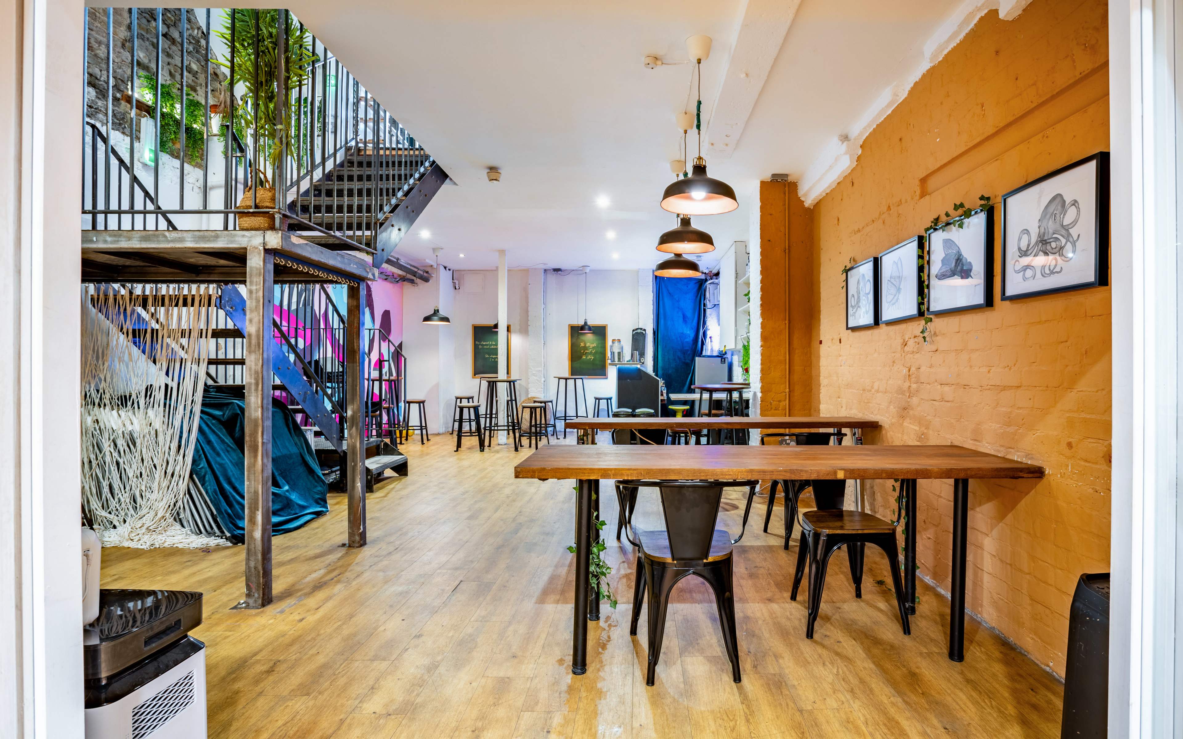 A spacious interior with wooden floors, metal tables, and an open stairway, featuring art on the walls and various seating arrangements.