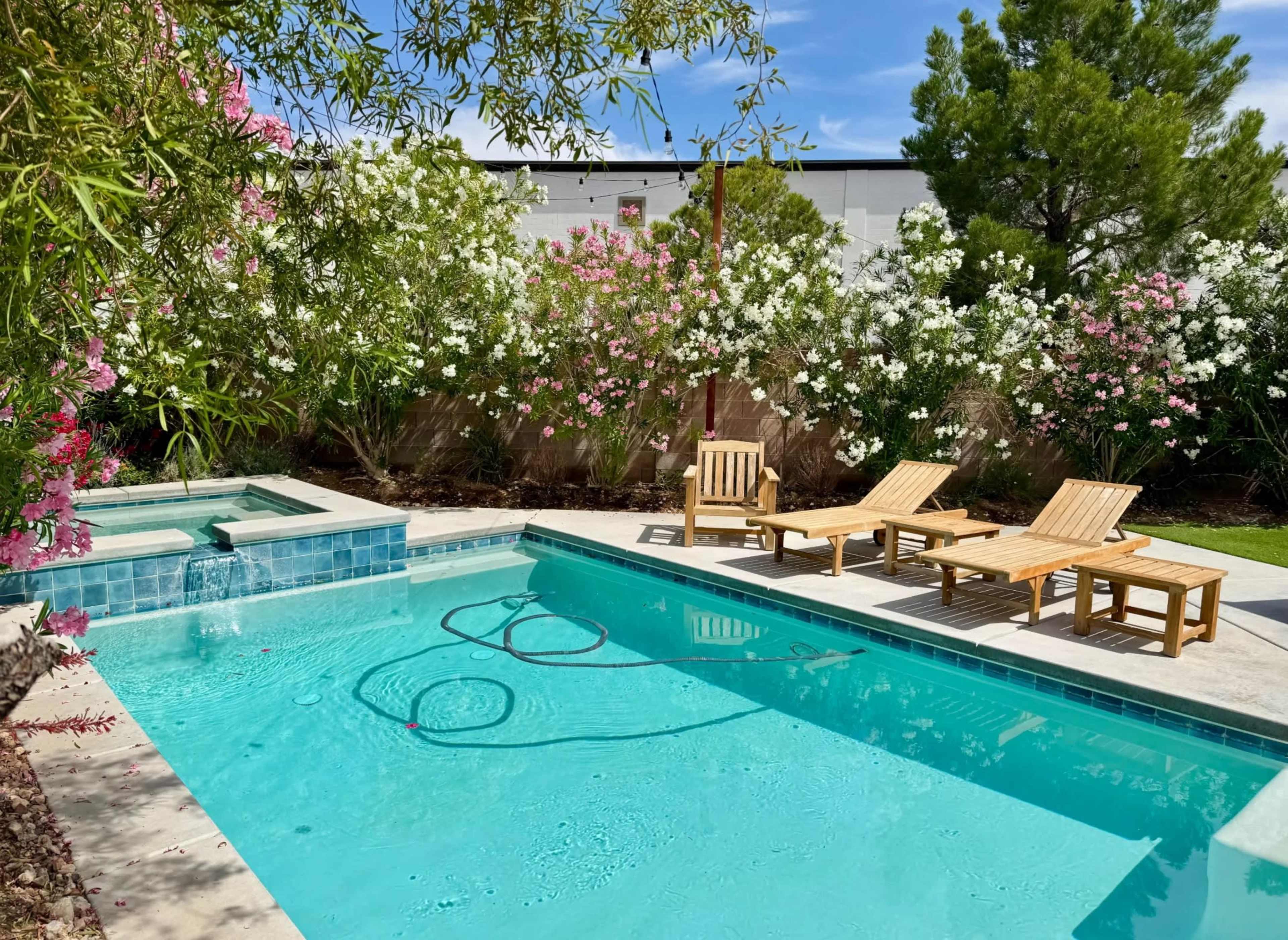 A clear swimming pool with a spa section, surrounded by blooming flowers and wooden lounge chairs under a blue sky.