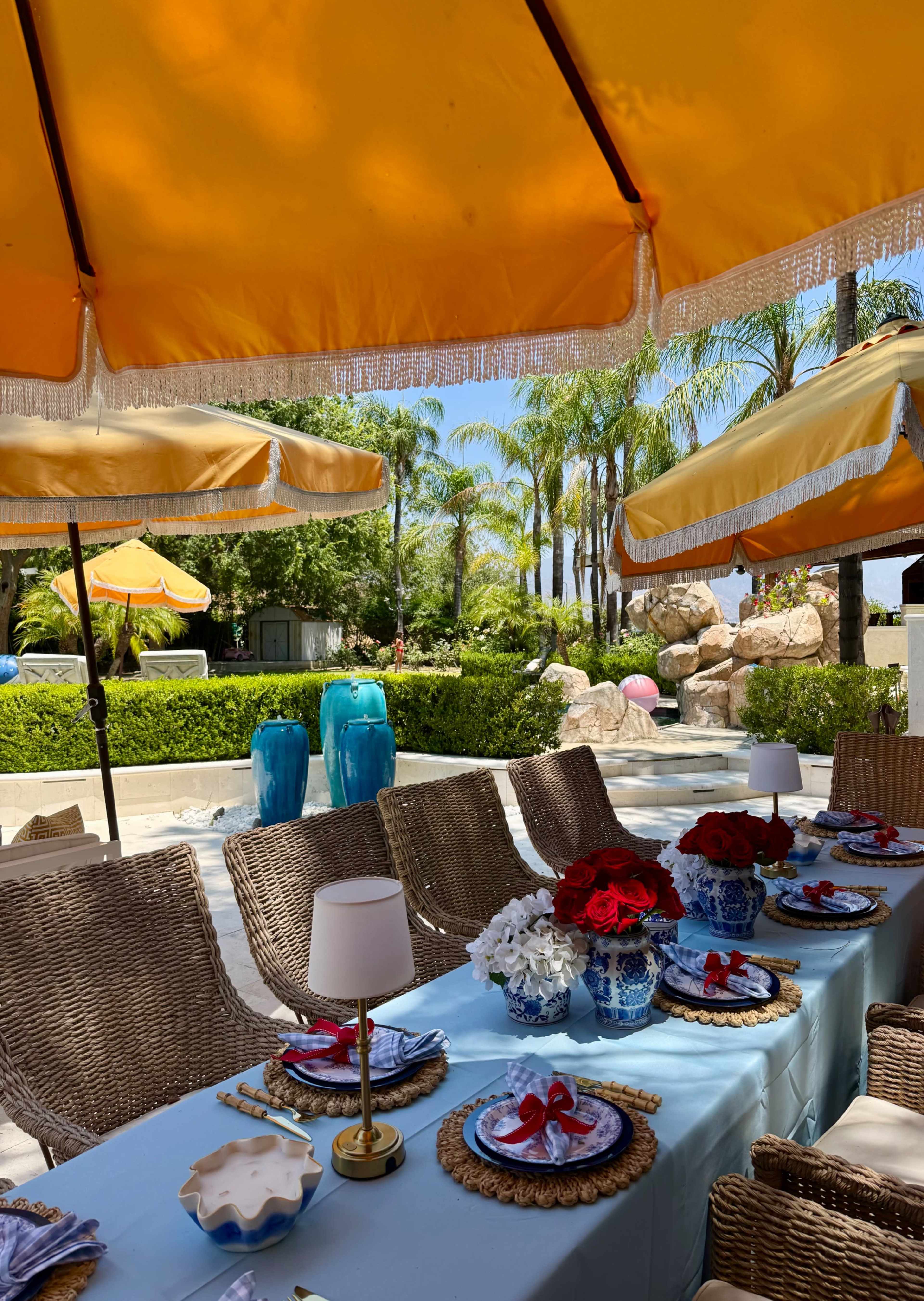 A neatly arranged outdoor dining table is set under orange umbrellas, surrounded by palm trees and a pool area.