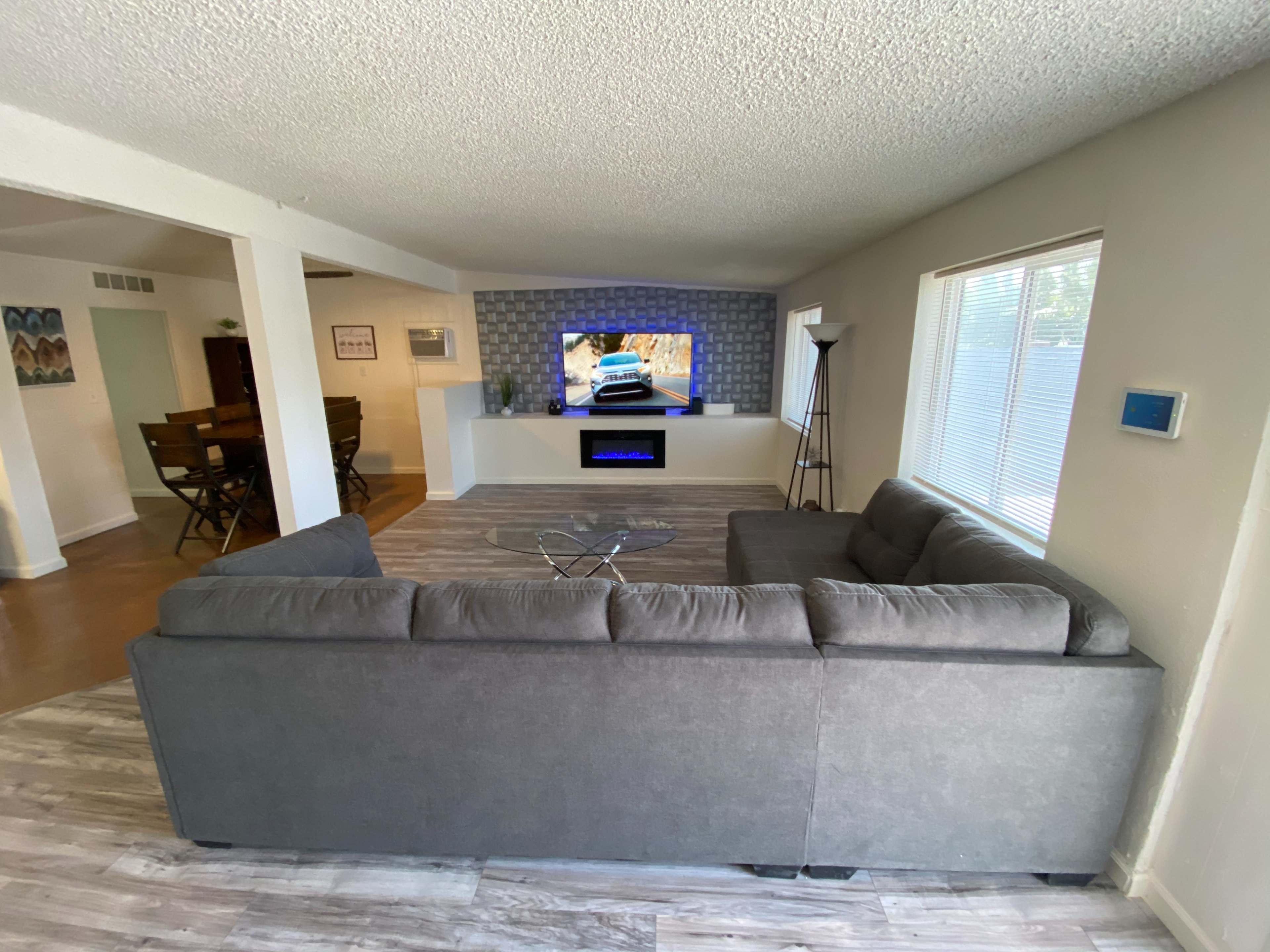 A modern living room features a large gray sectional sofa facing a wall-mounted television and an electric fireplace, with a dining area visible in the background.
