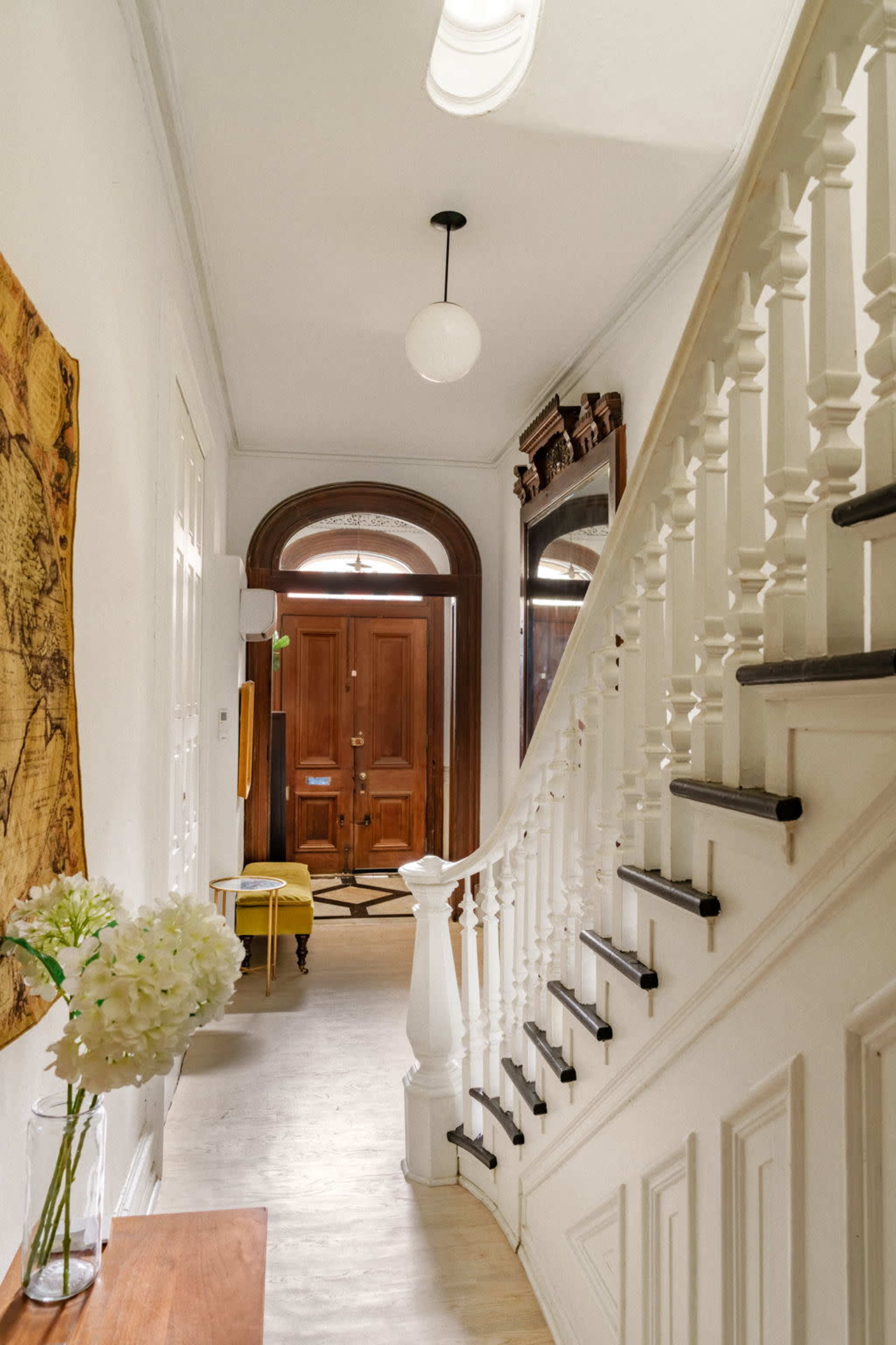 The image shows a well-lit hallway featuring a wooden staircase, a decorative mirror, and a door with a round window at the entrance.