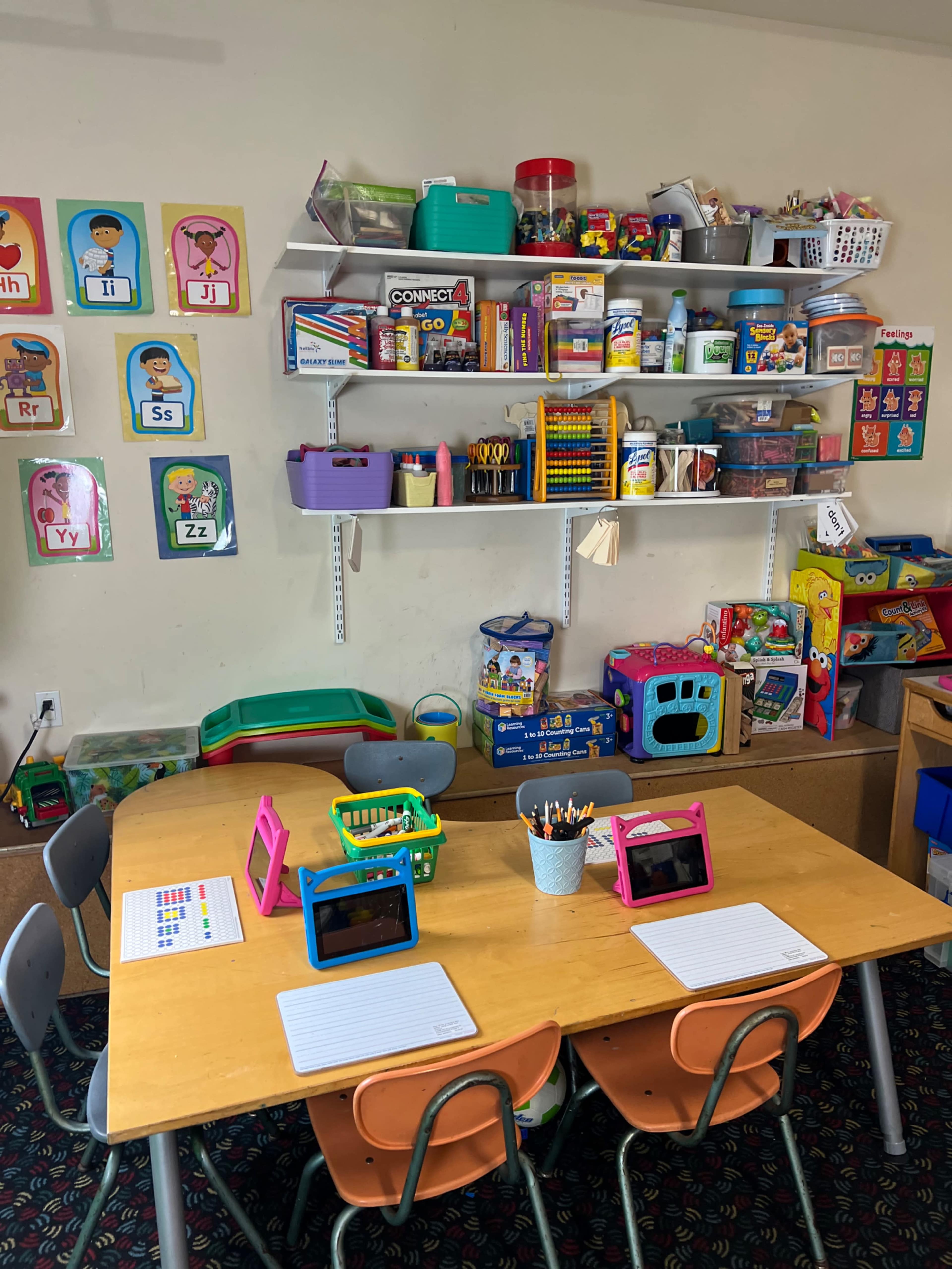 A classroom space features two tables with pink and green tablet holders, surrounded by shelves filled with educational materials and colorful toys.