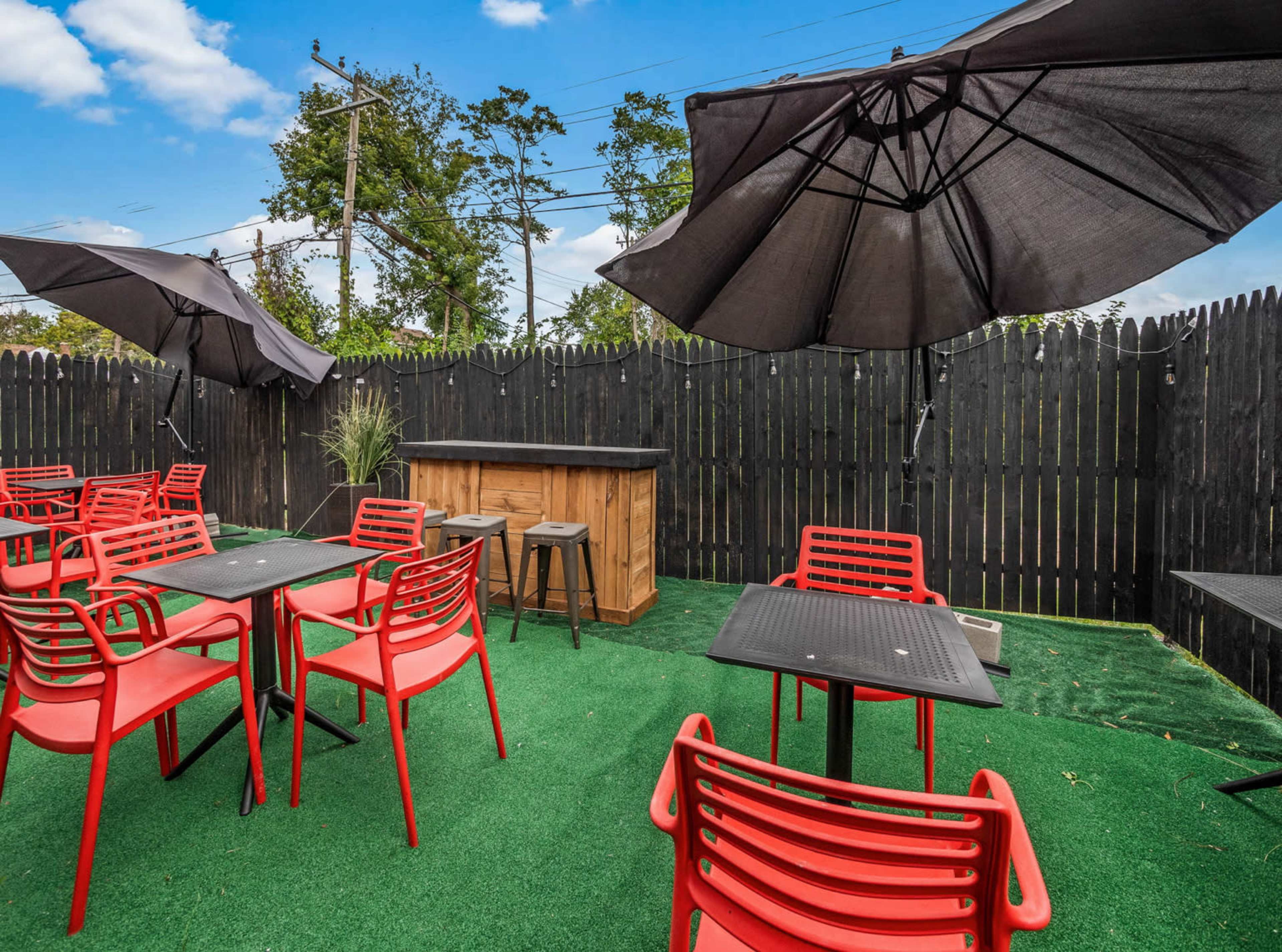 The image shows an outdoor seating area with red chairs and tables, featuring large black umbrellas and a small wooden bar against a backdrop of a black wooden fence.