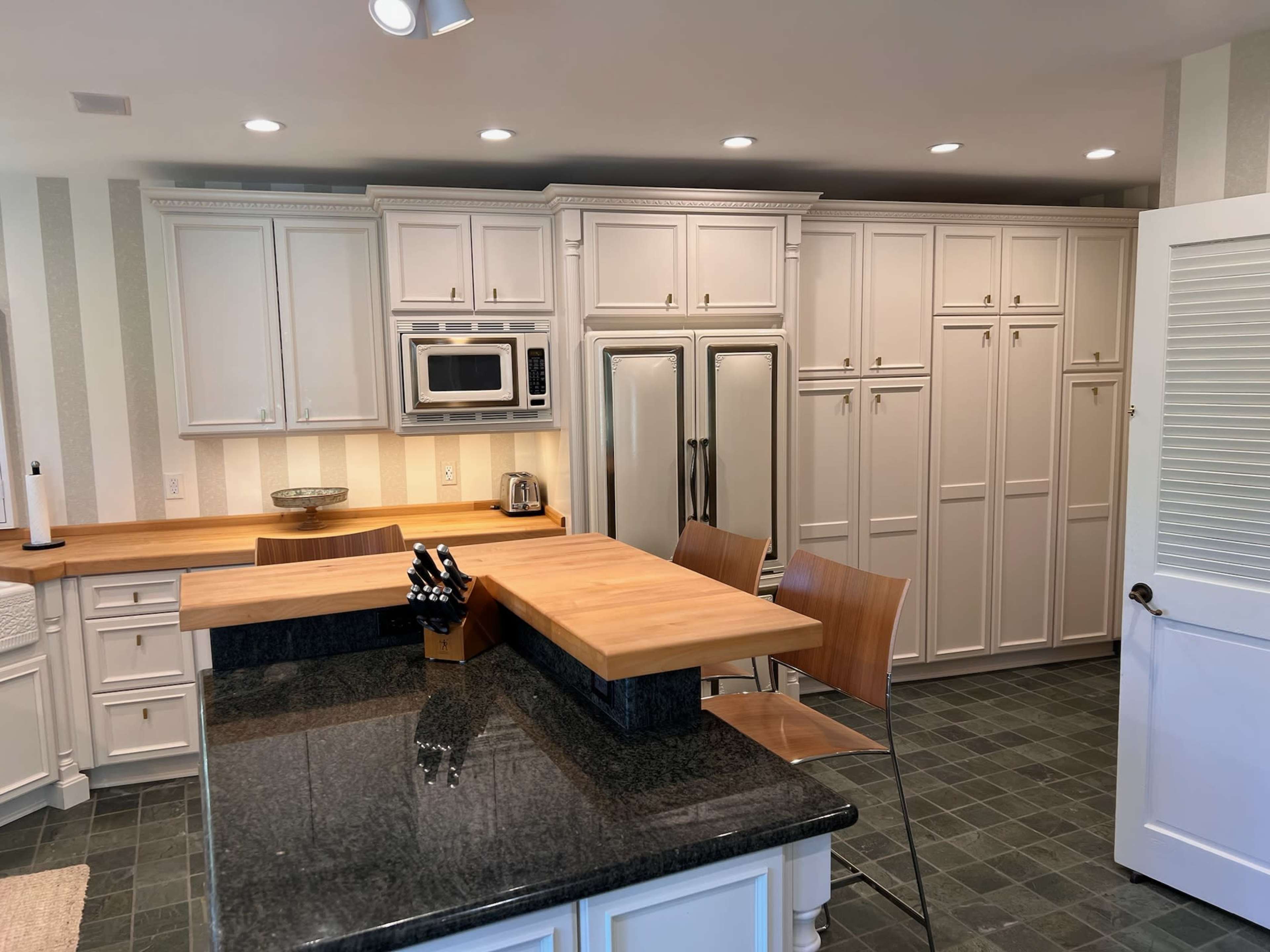 The image shows a modern kitchen with white cabinetry, a dark granite countertop, and wooden bar stools at a breakfast bar.