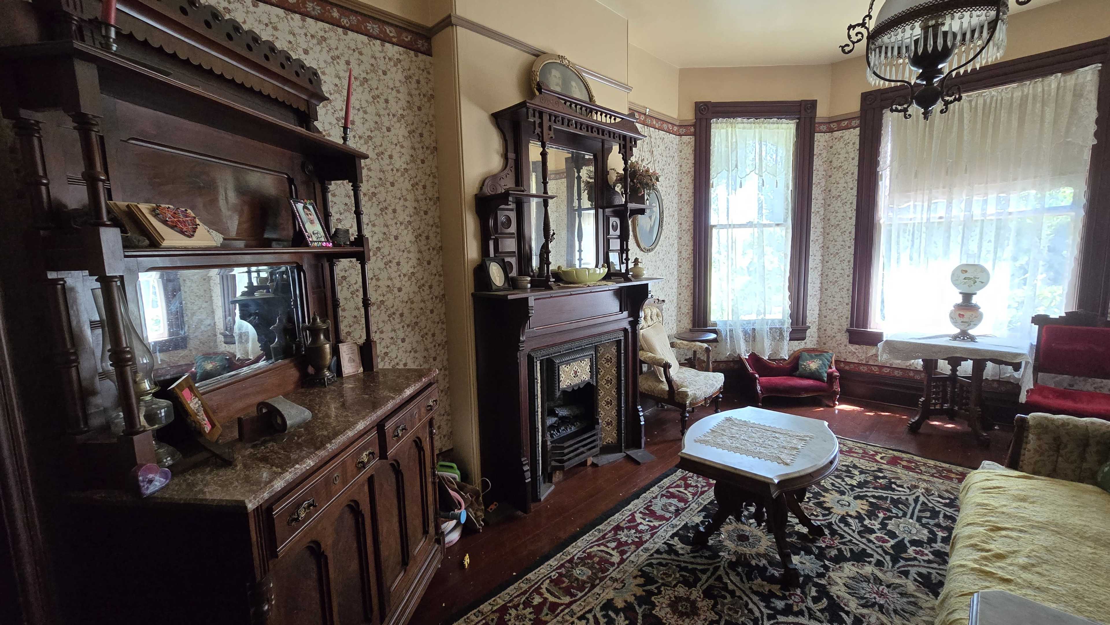 The room features ornate wooden furniture, a fireplace, and large windows with sheer curtains, all set against patterned wallpaper and a colorful rug.