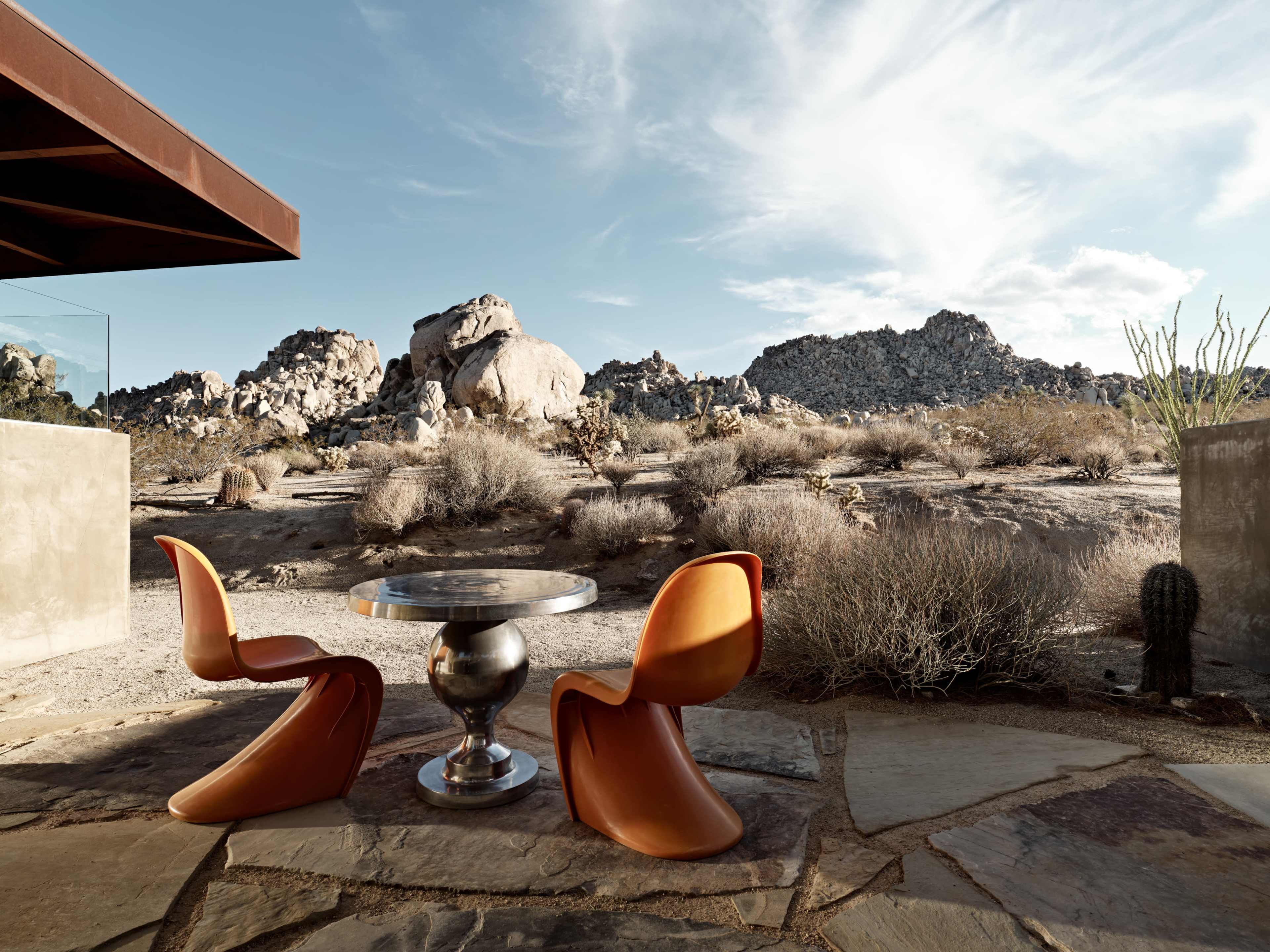 A modern outdoor dining area features a round table and two orange chairs, set against a backdrop of rocky desert terrain and clear blue skies.