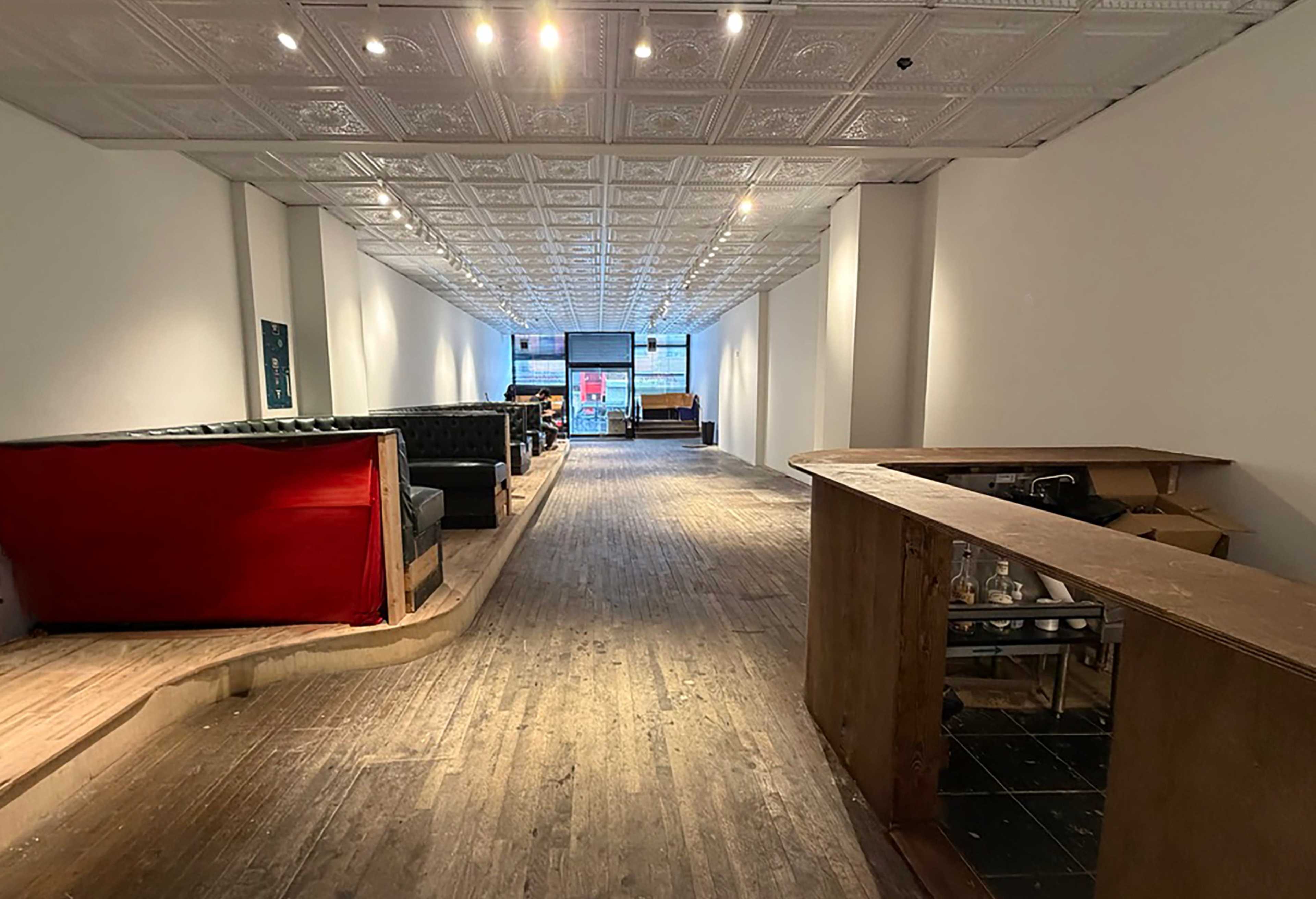 The image shows a long, narrow room with hardwood floors, a bar counter on the right, and scattered seating along the walls, illuminated by overhead lights.