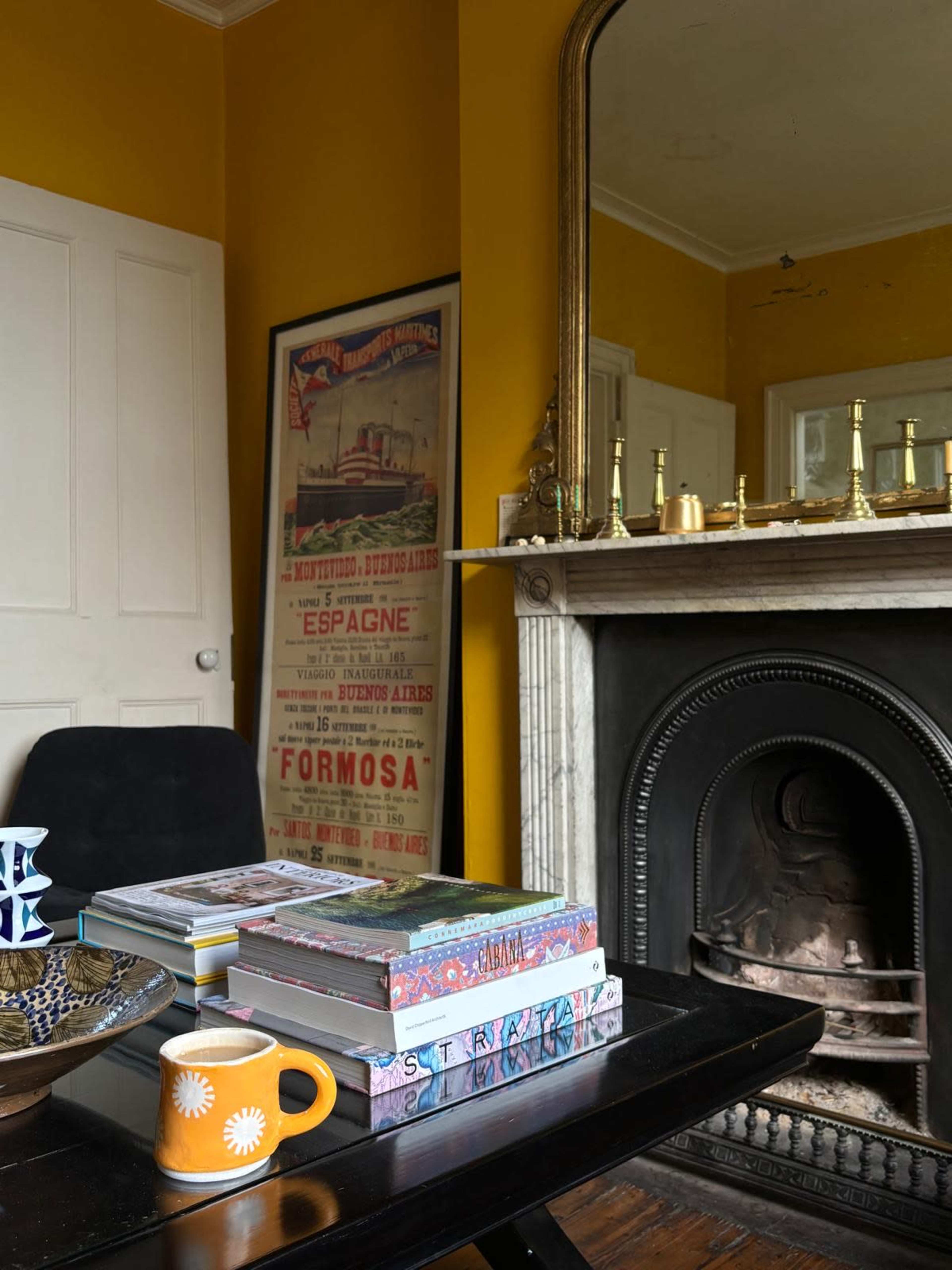 A cozy room features a black chair and a coffee table stacked with books, alongside a decorative bowl and a colorful mug, all set against a vibrant yellow wall and a mirrored fireplace.