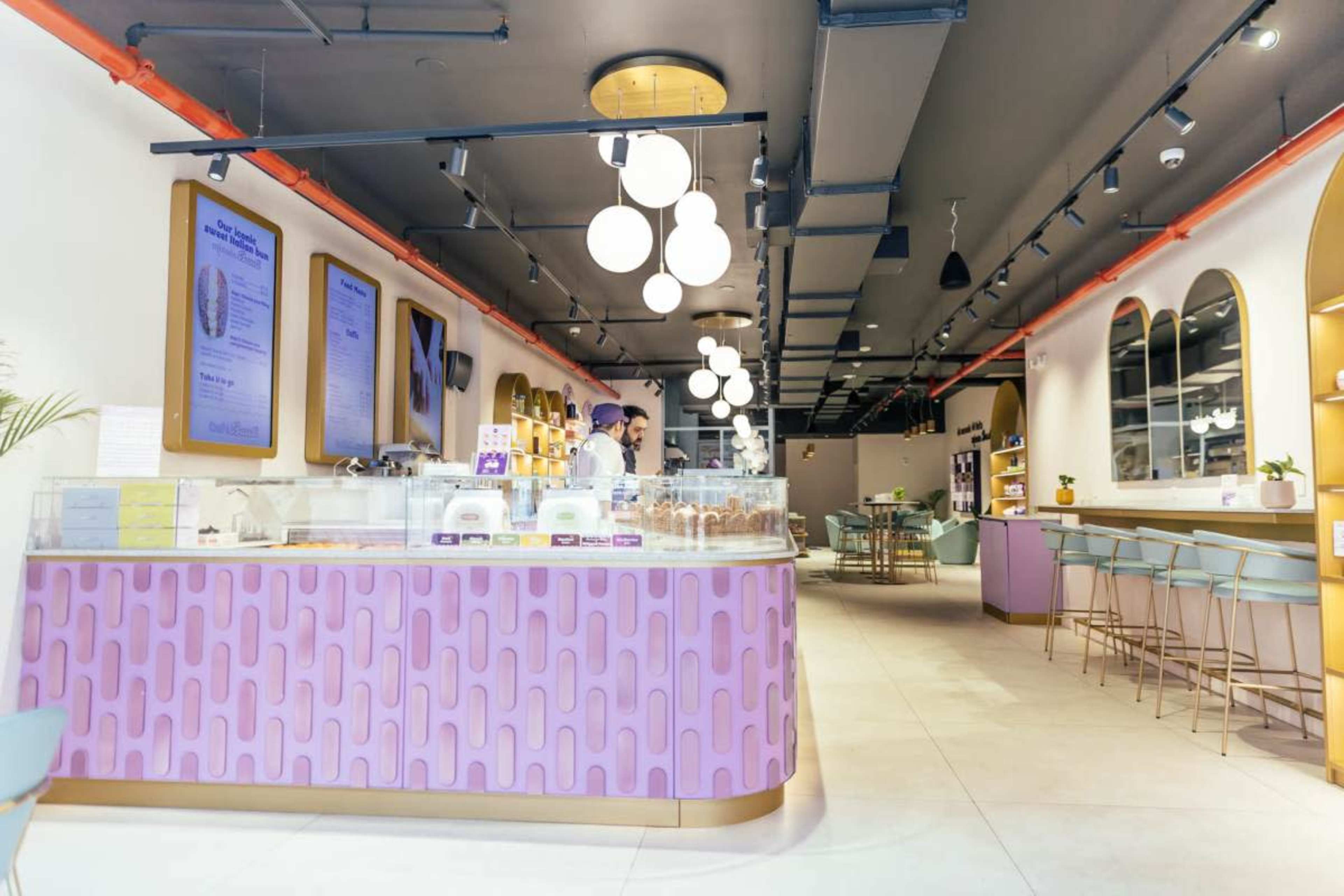 The image shows a modern bakery with a brightly colored counter displaying various desserts, surrounded by seating and stylish decor.