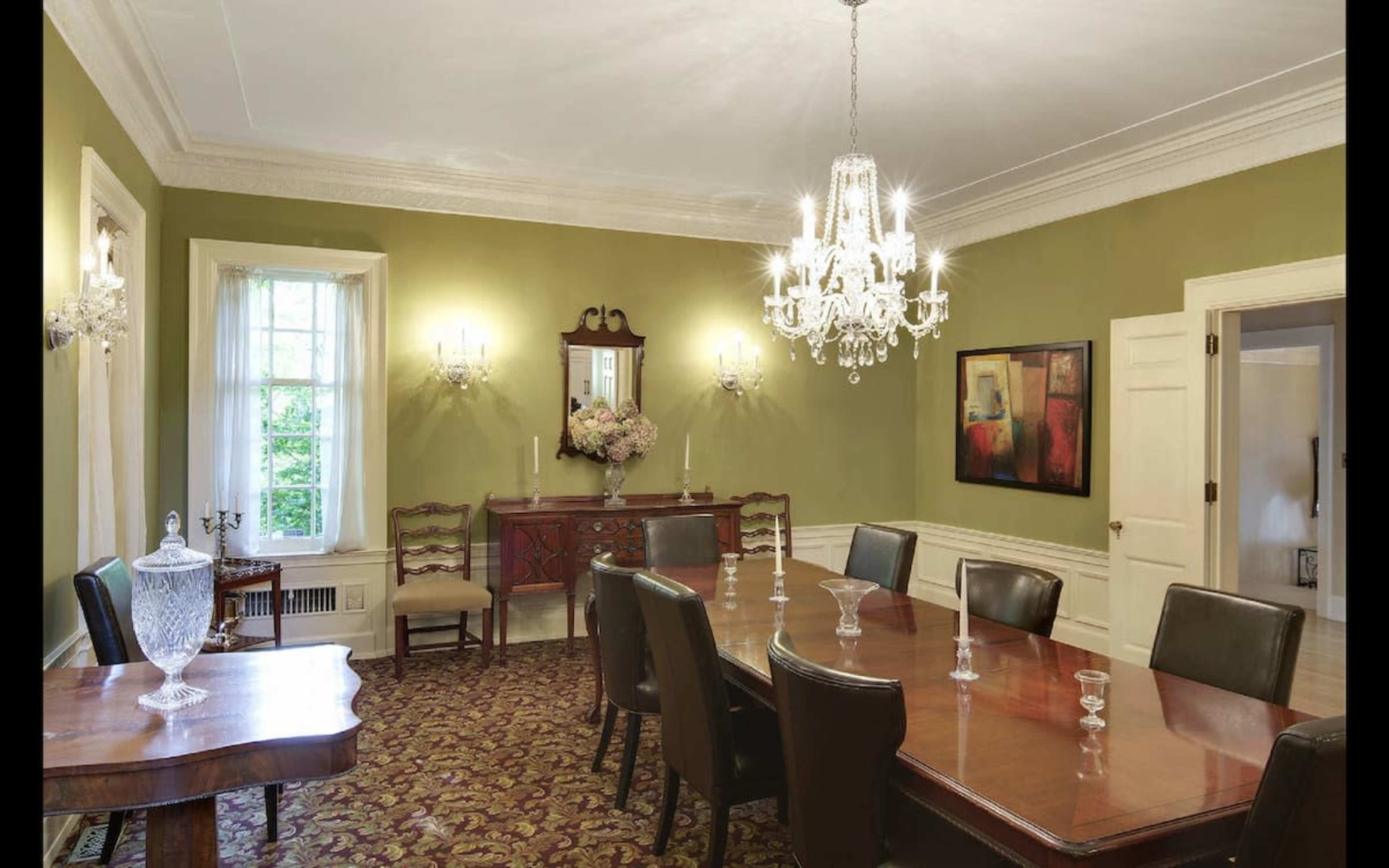 A well-furnished dining room featuring a long wooden table, chairs, a chandelier, and a sideboard against a green wall.