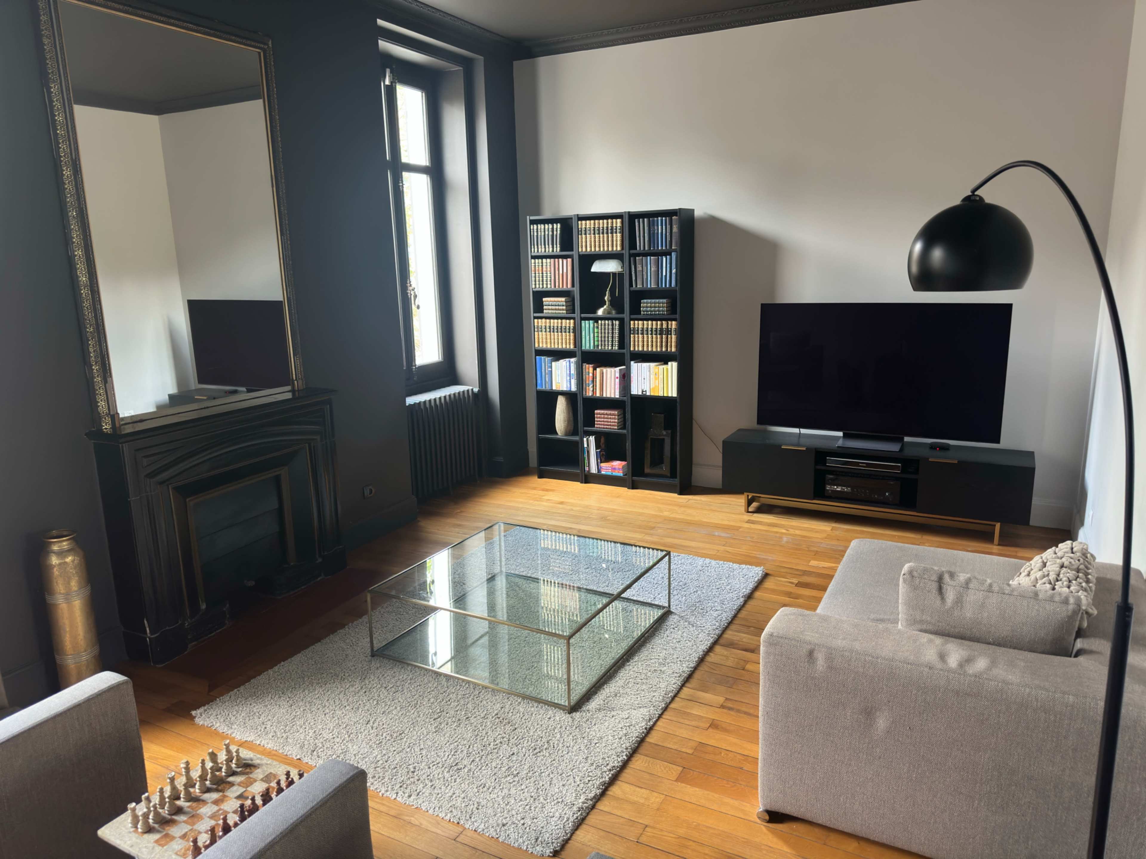 The image shows a modern living room with a glass coffee table, a grey couch, a bookshelf filled with books, a television stand, and a large mirror on one wall.