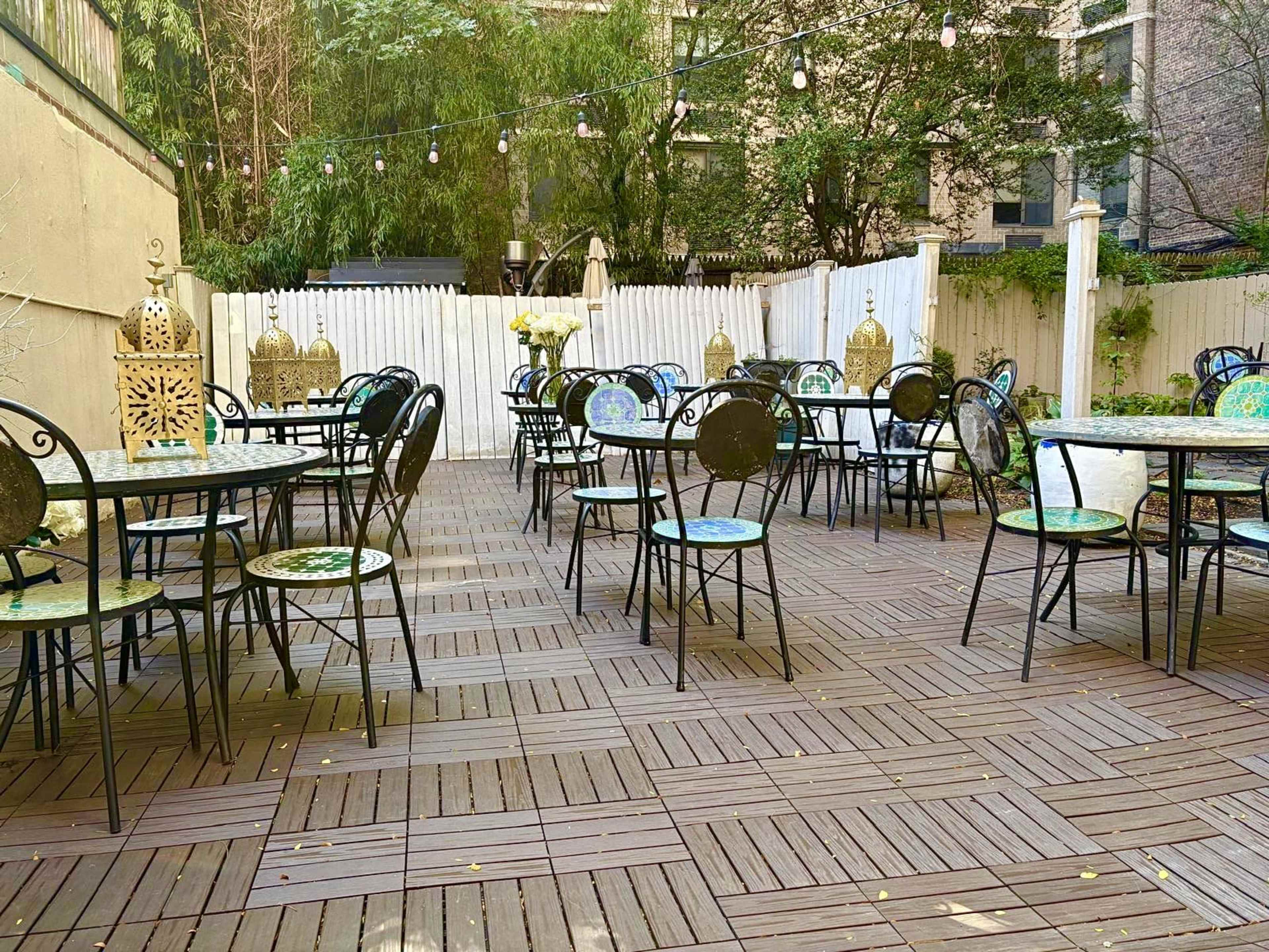 The image shows an outdoor dining area with several round tables and black metal chairs arranged on a wooden floor, surrounded by greenery and decorative lanterns.