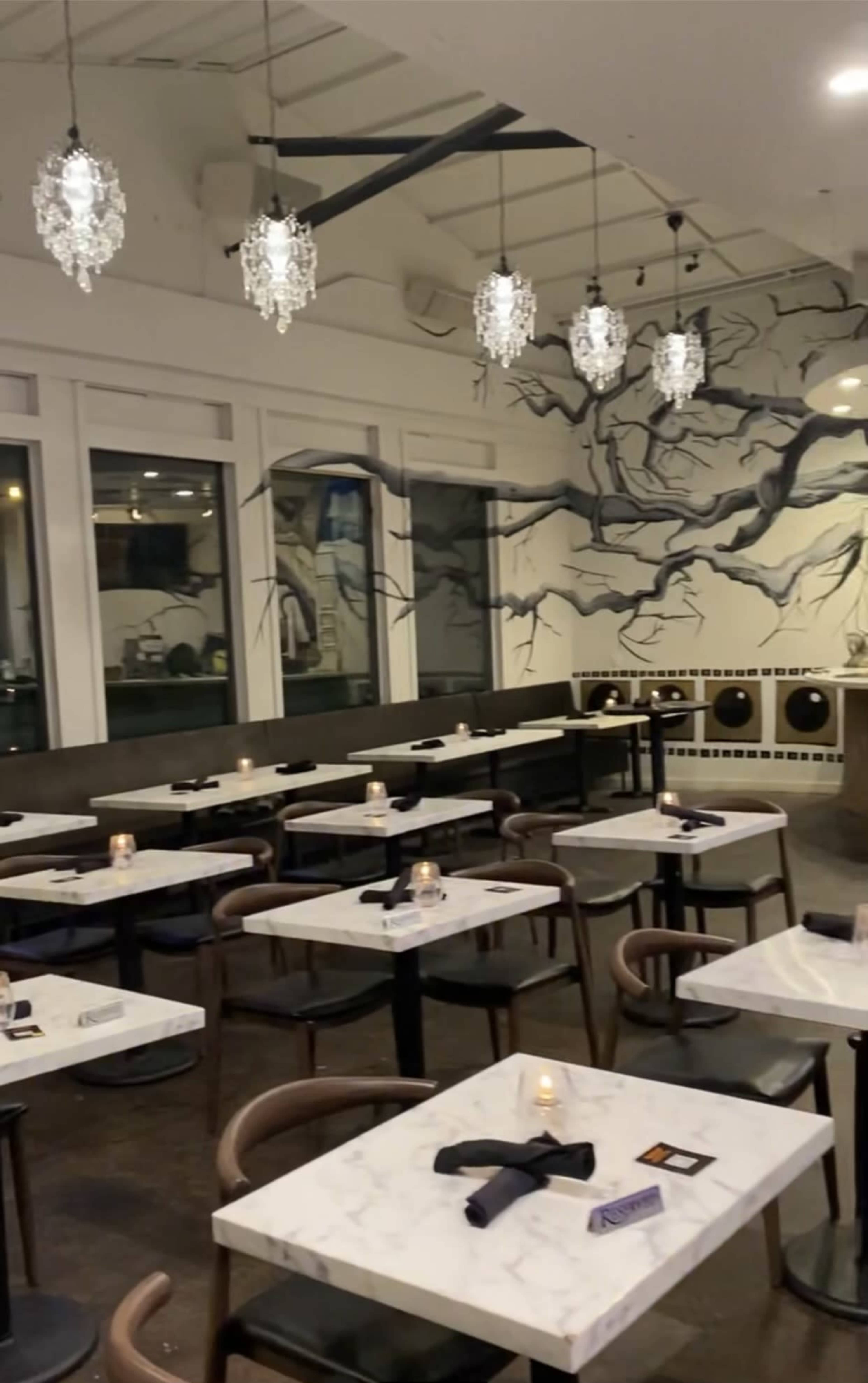 A modern restaurant interior with marble-topped tables, dark chairs, and a wall adorned with an artistic tree mural, illuminated by hanging chandeliers.