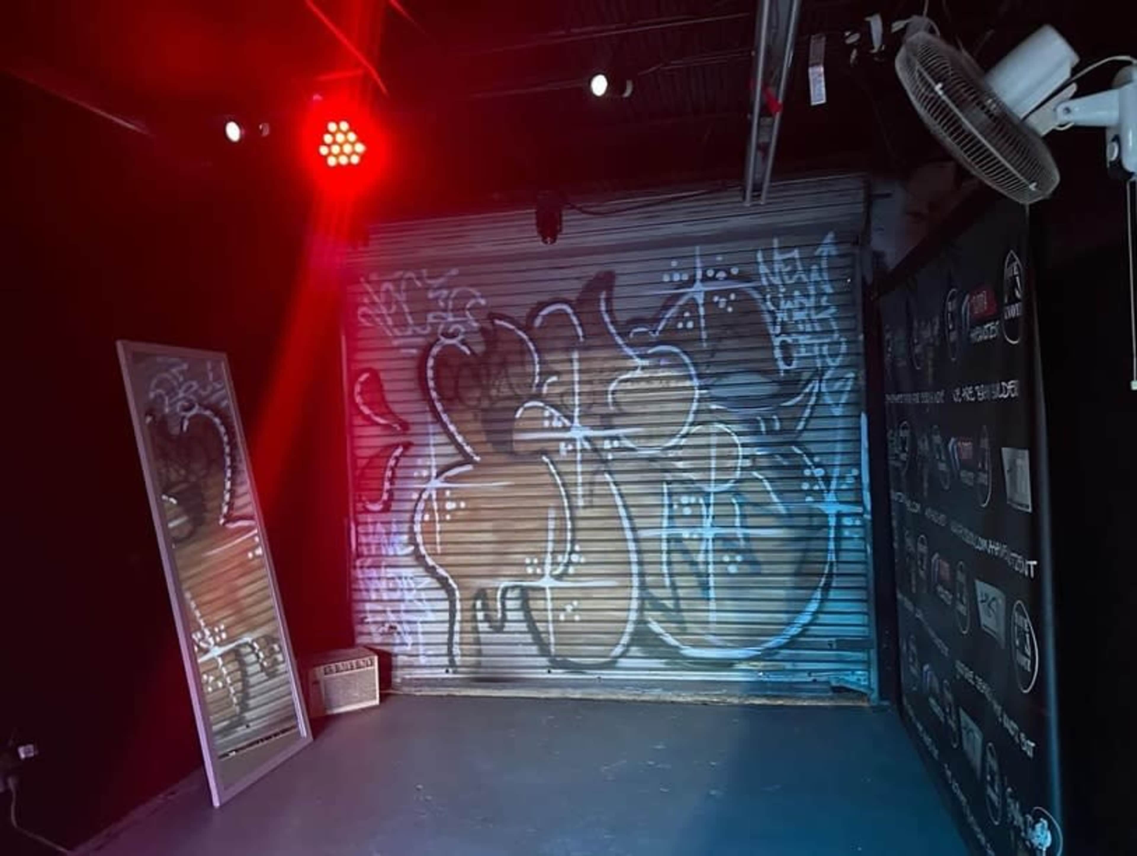 The image shows a corner of a room with a graffiti-covered metal shutter and a large mirror, illuminated by red lighting.