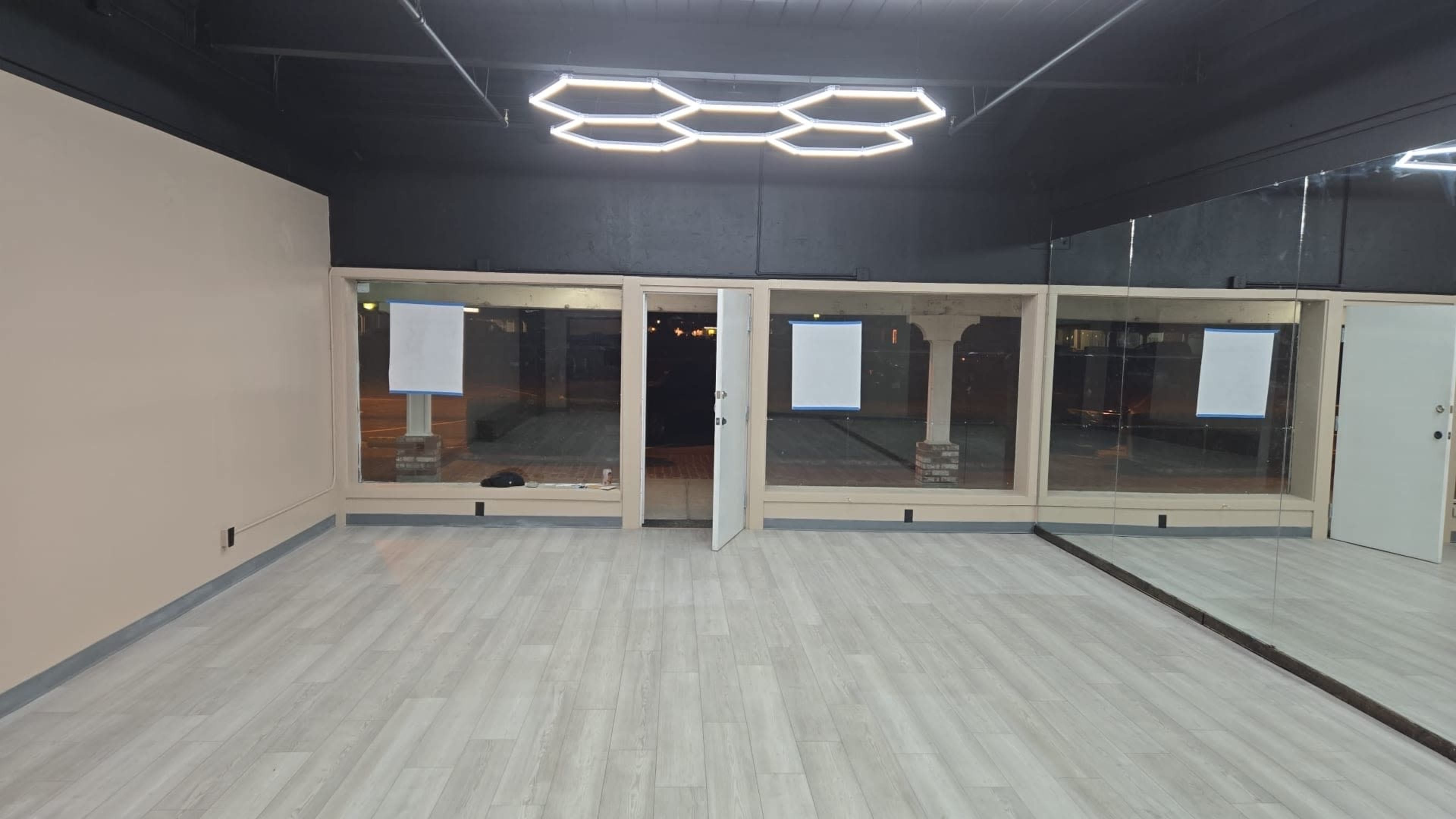 The image shows an empty room with a light-colored floor and two large windowed sections, illuminated by a modern hexagonal light fixture above.