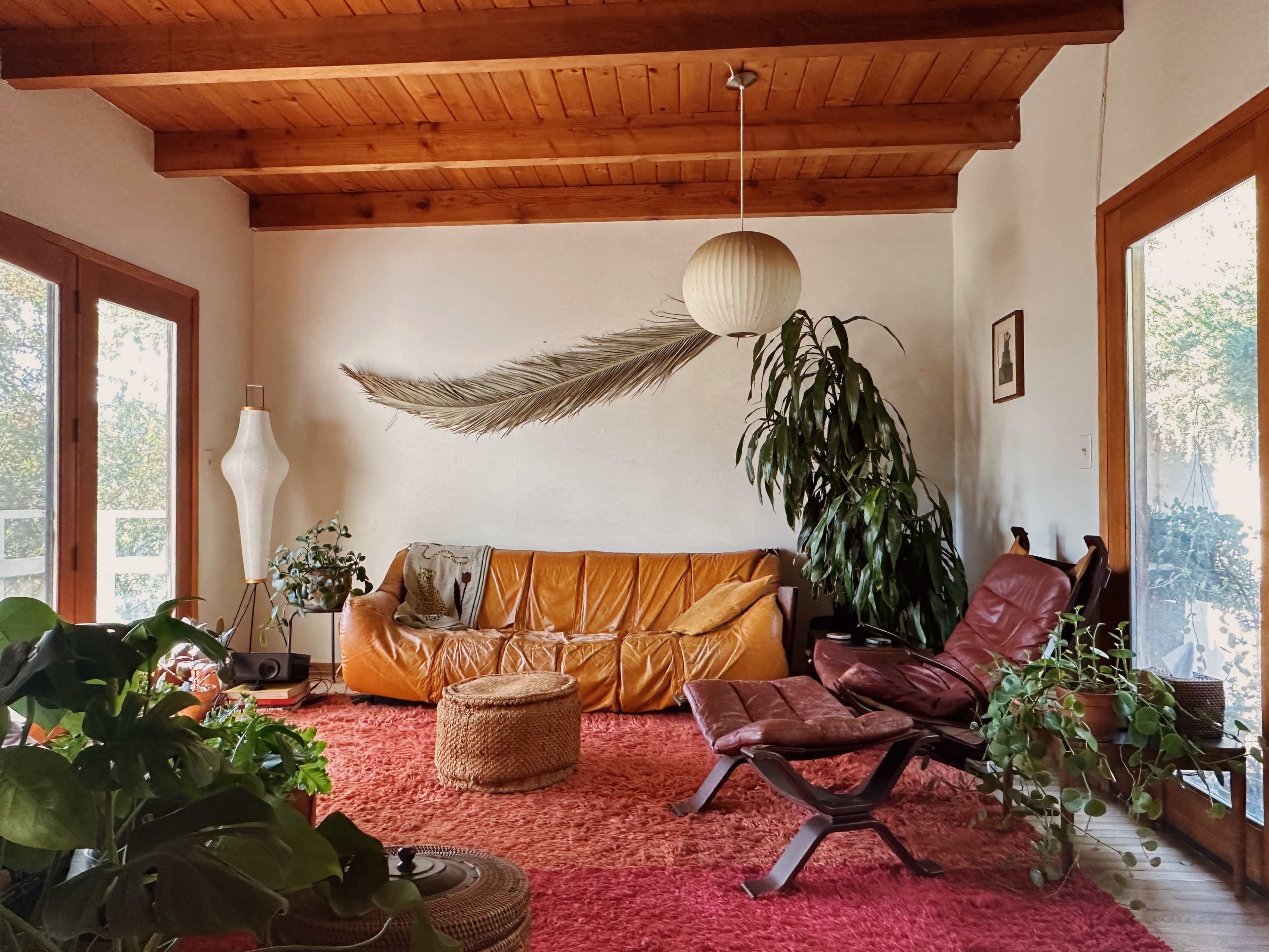 The room features a brown leather sofa, a red rug, a woven ottoman, and large windows surrounded by greenery.