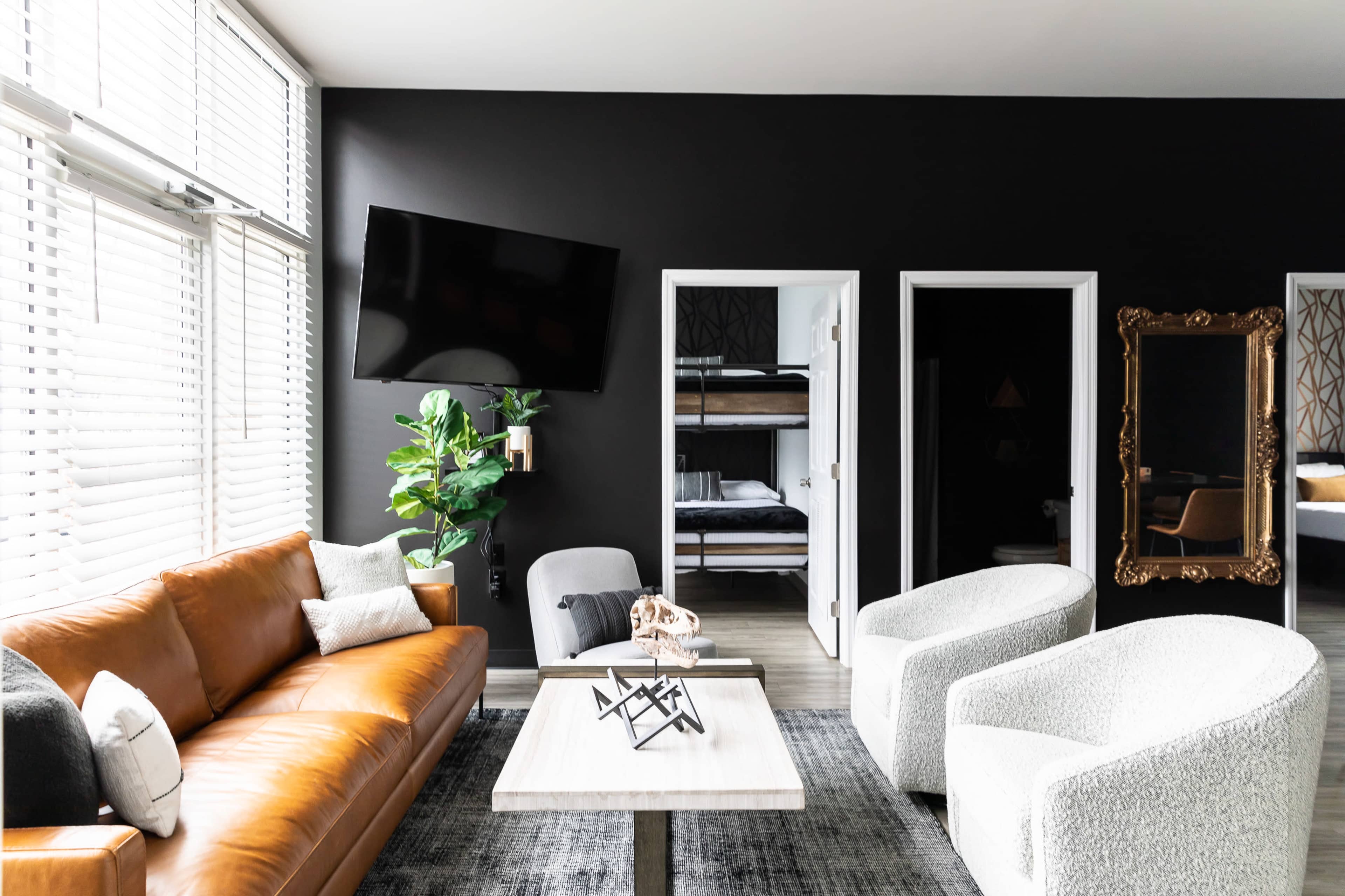A modern living room features a brown leather sofa, two white armchairs, a wooden coffee table, and a large television mounted on a dark wall, with a potted plant beside a doorway leading to a bunk bedroom.