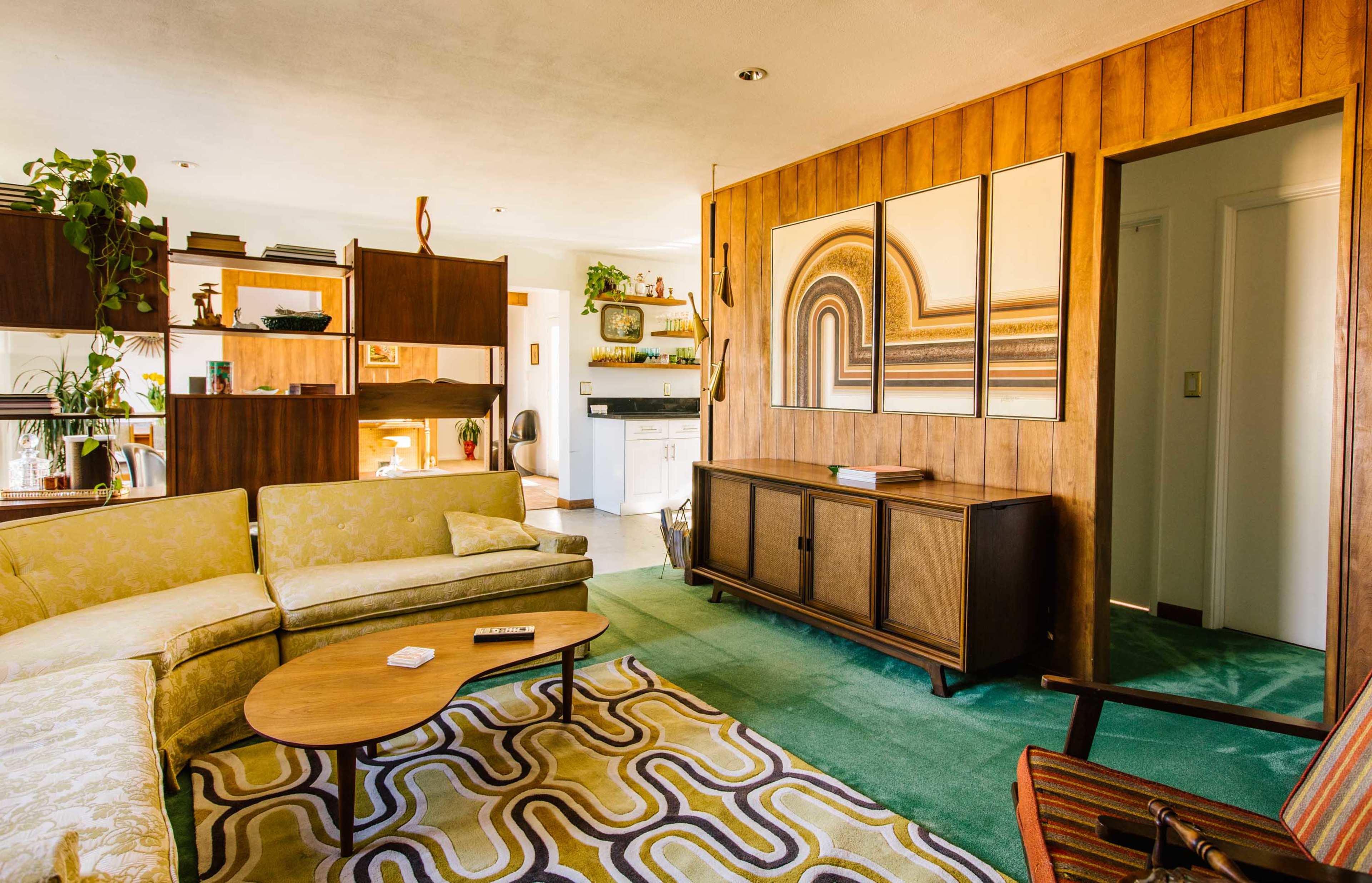 A mid-century modern living room featuring a green carpet, a patterned area rug, a sectional sofa, and framed artwork on the wooden-paneled wall.