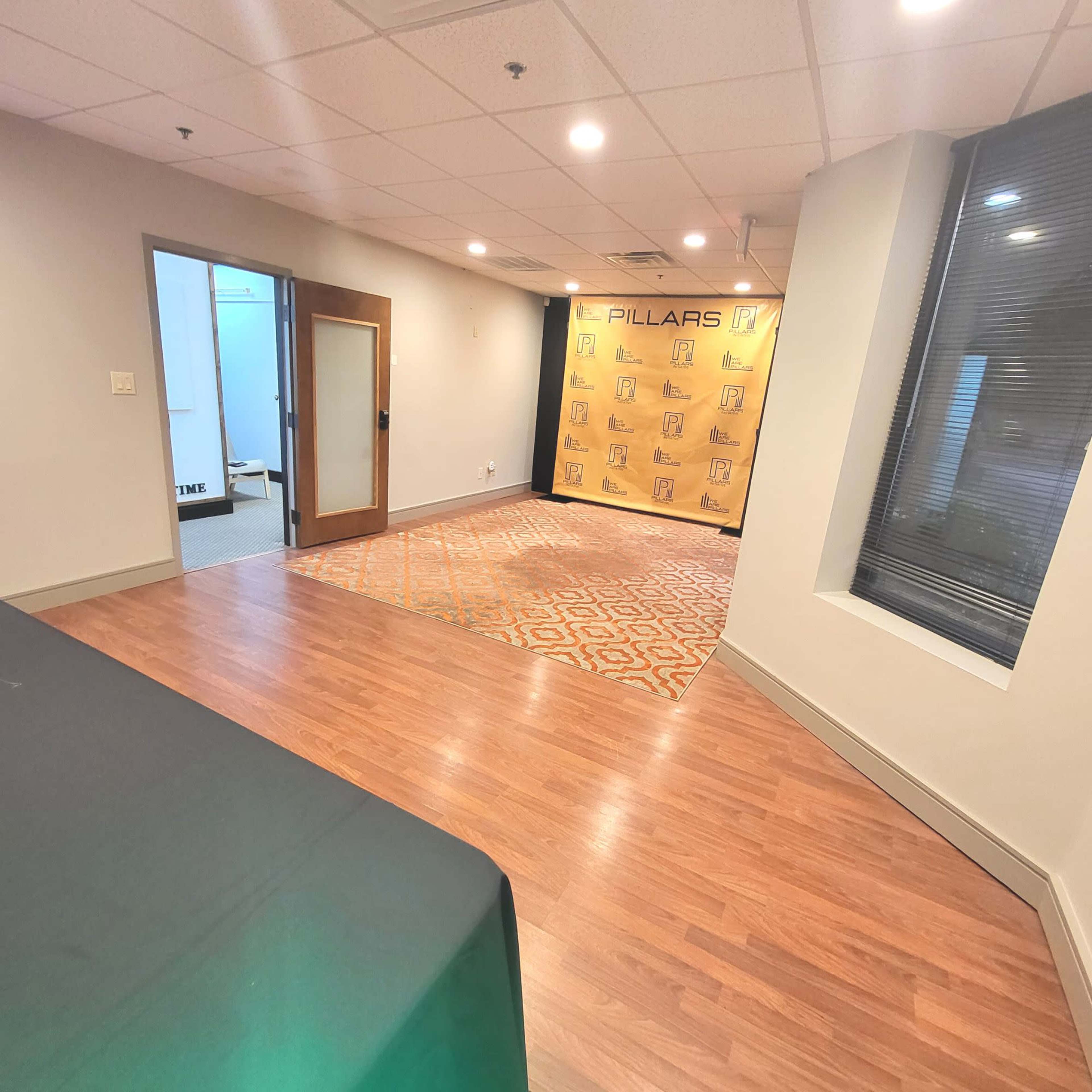 A brightly lit room with wooden flooring, a patterned rug, and a backdrop featuring the word "PILLARS" on a wall.
