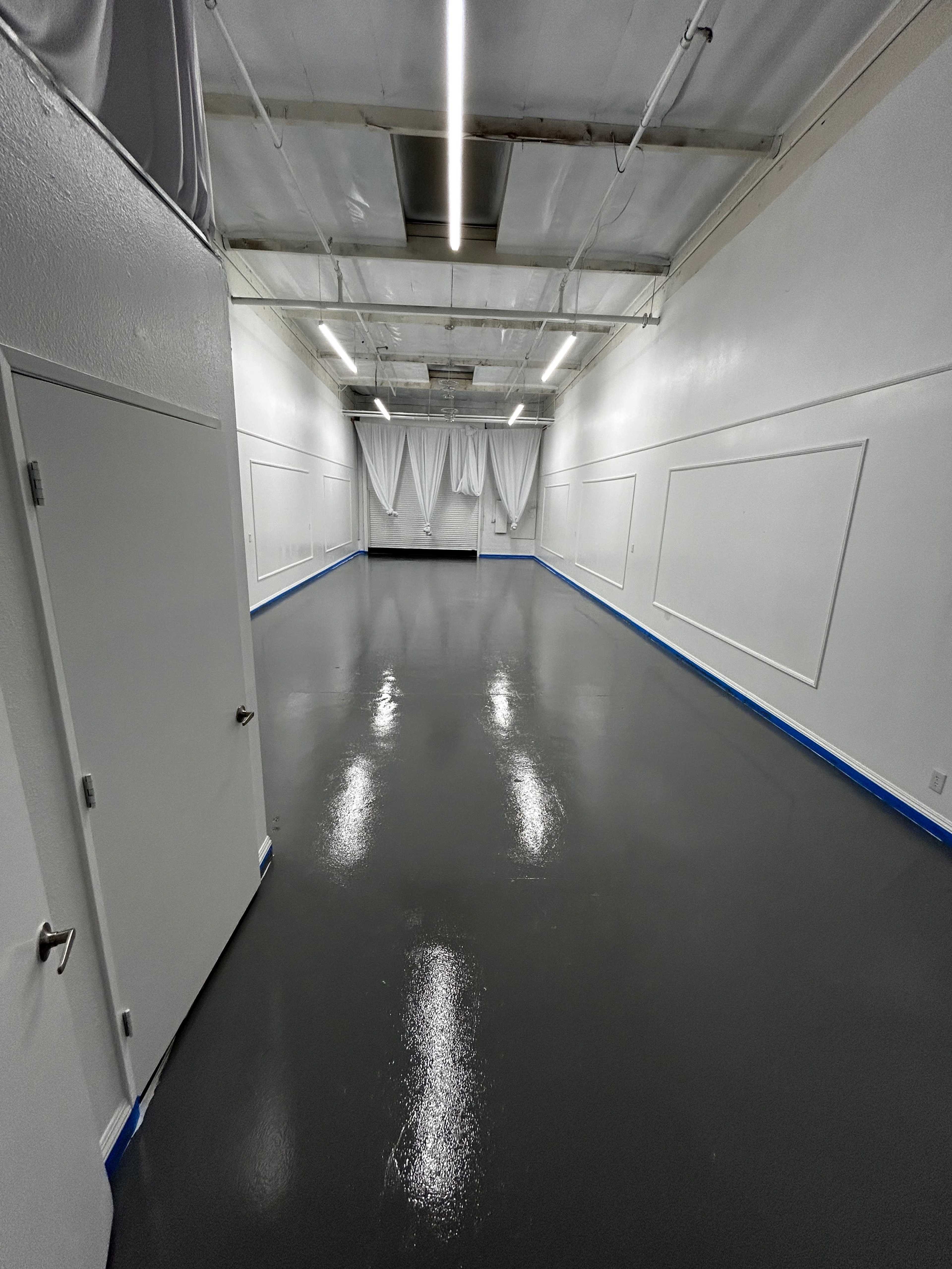 A spacious, empty room with a polished gray floor and white walls, featuring two doors and curtains at one end.