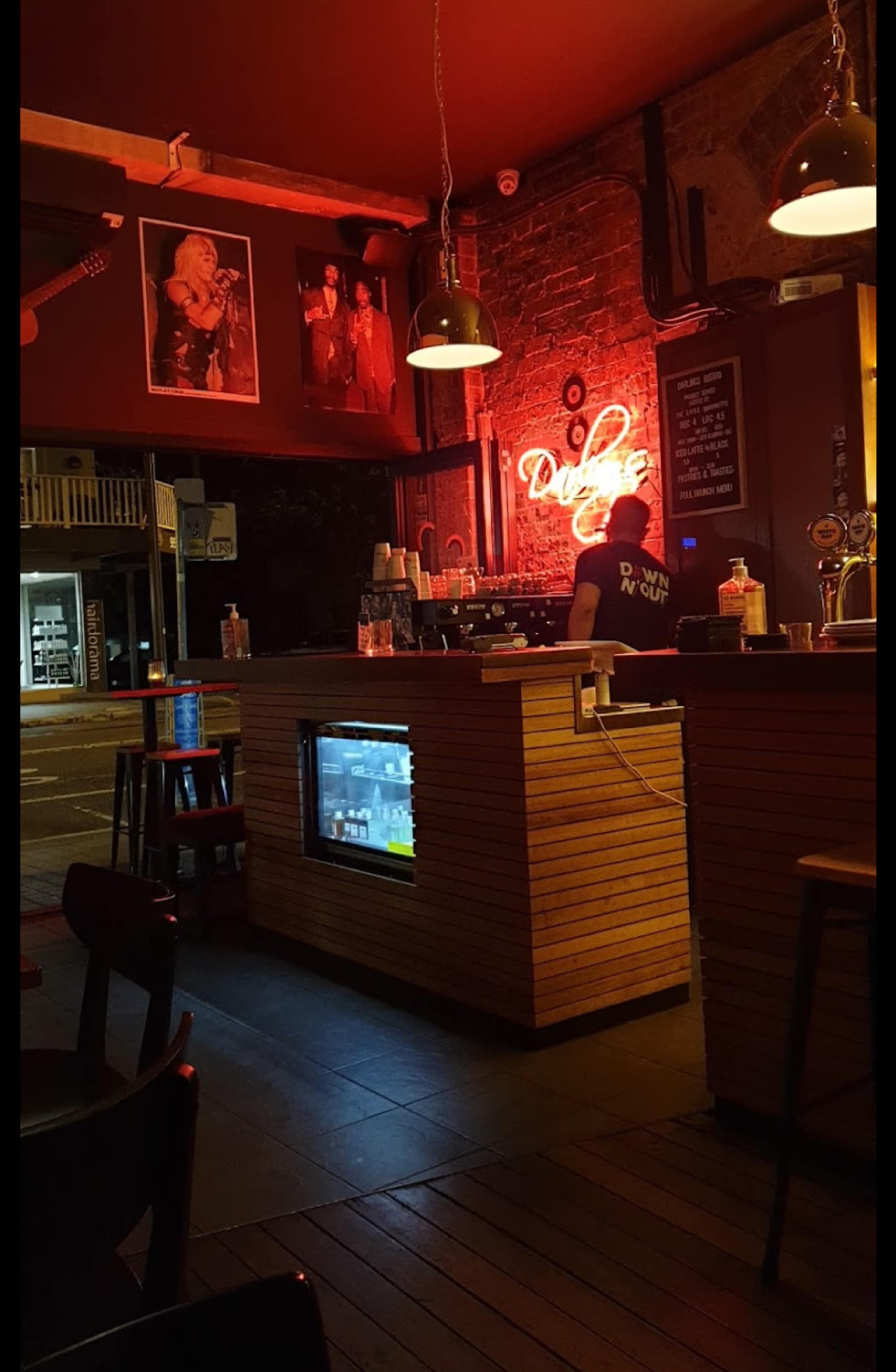 The image shows the interior of a dimly lit bar with a wooden counter, neon signage, and a bartender preparing drinks against a backdrop of framed photographs on the wall.