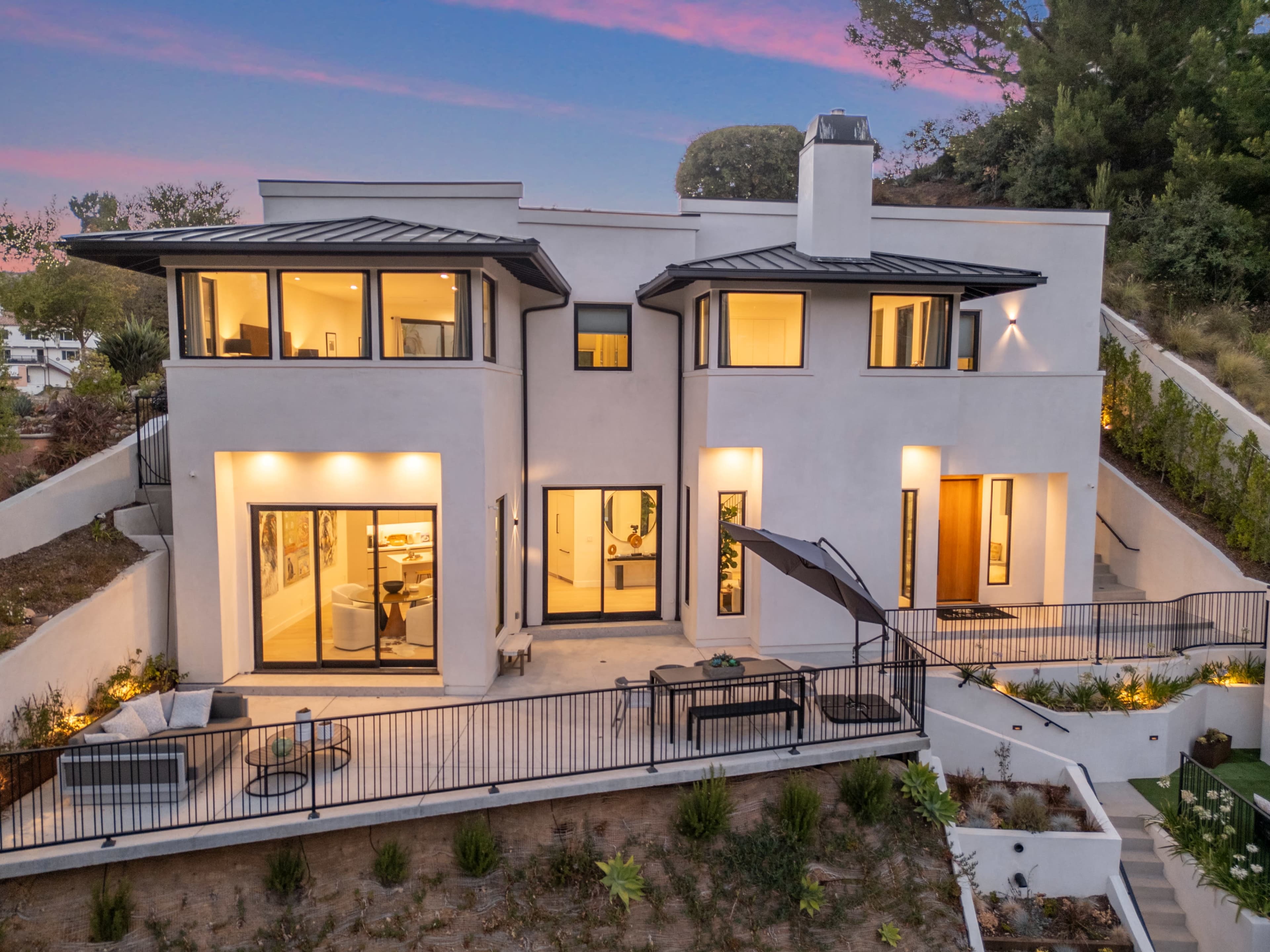 A modern two-story house with large windows and a balcony overlooks a landscaped hillside.