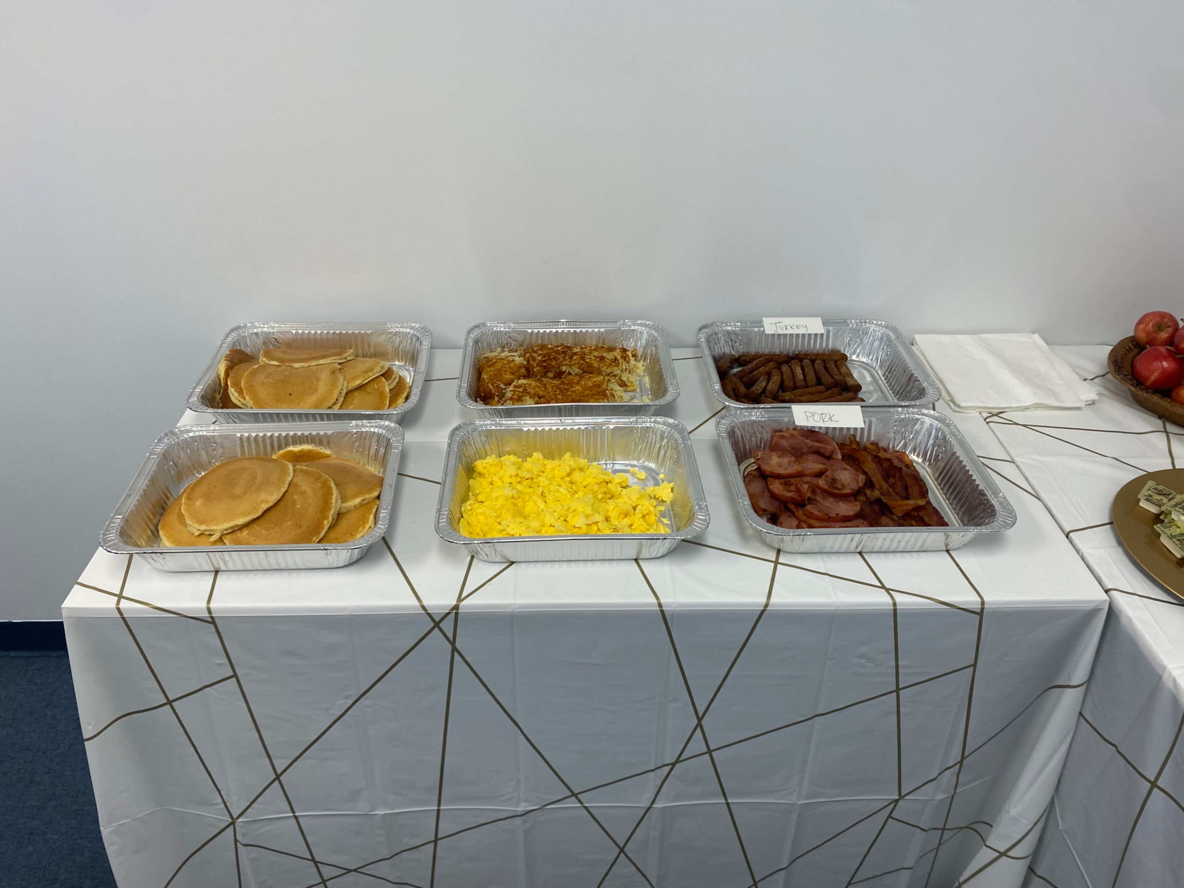 The image shows a table covered with a white tablecloth featuring six trays of breakfast food, including pancakes, scrambled eggs, sausage links, bacon, and a baked dish.