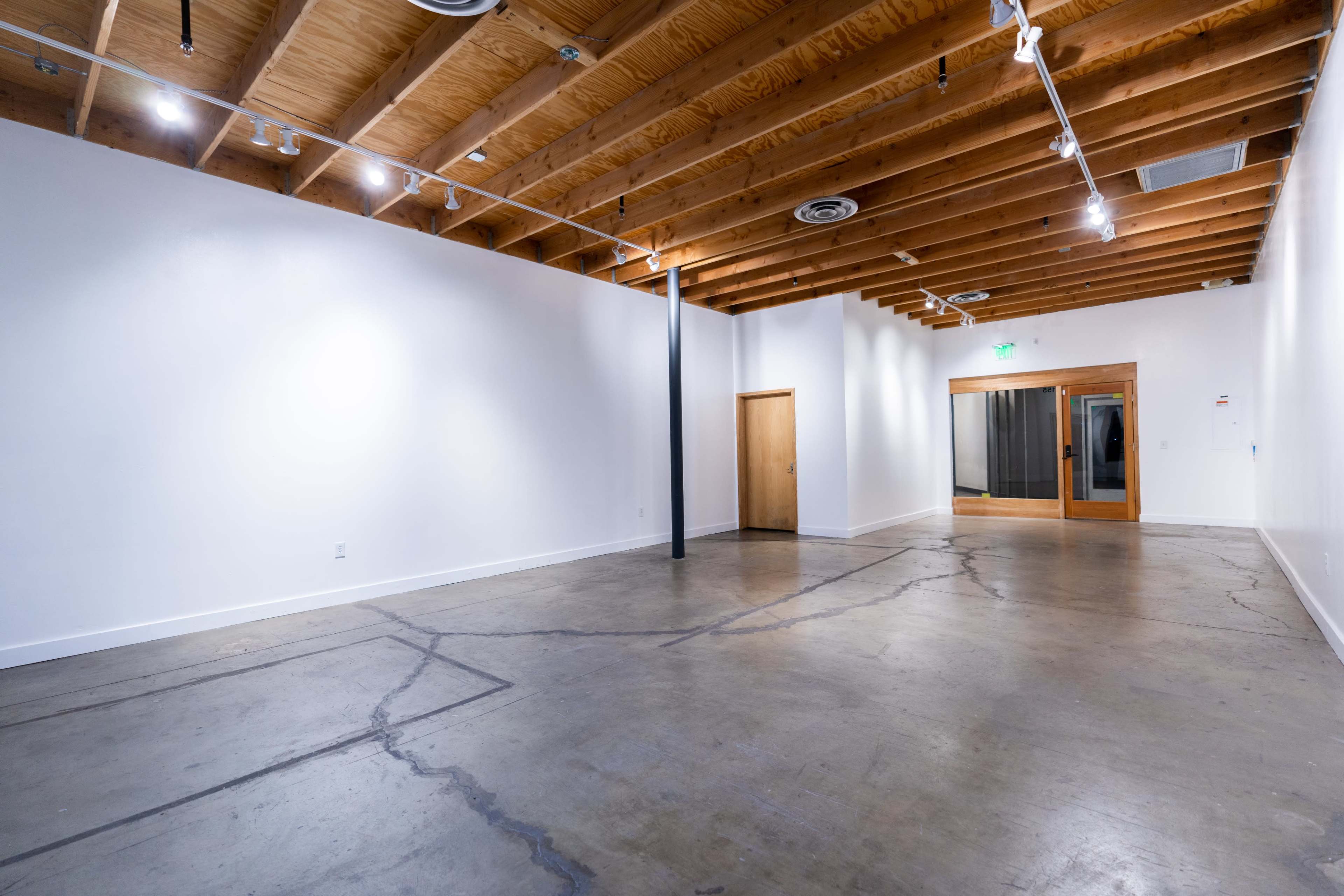 An empty gallery space with a polished concrete floor, wooden beams on the ceiling, and a doorway leading to another room.