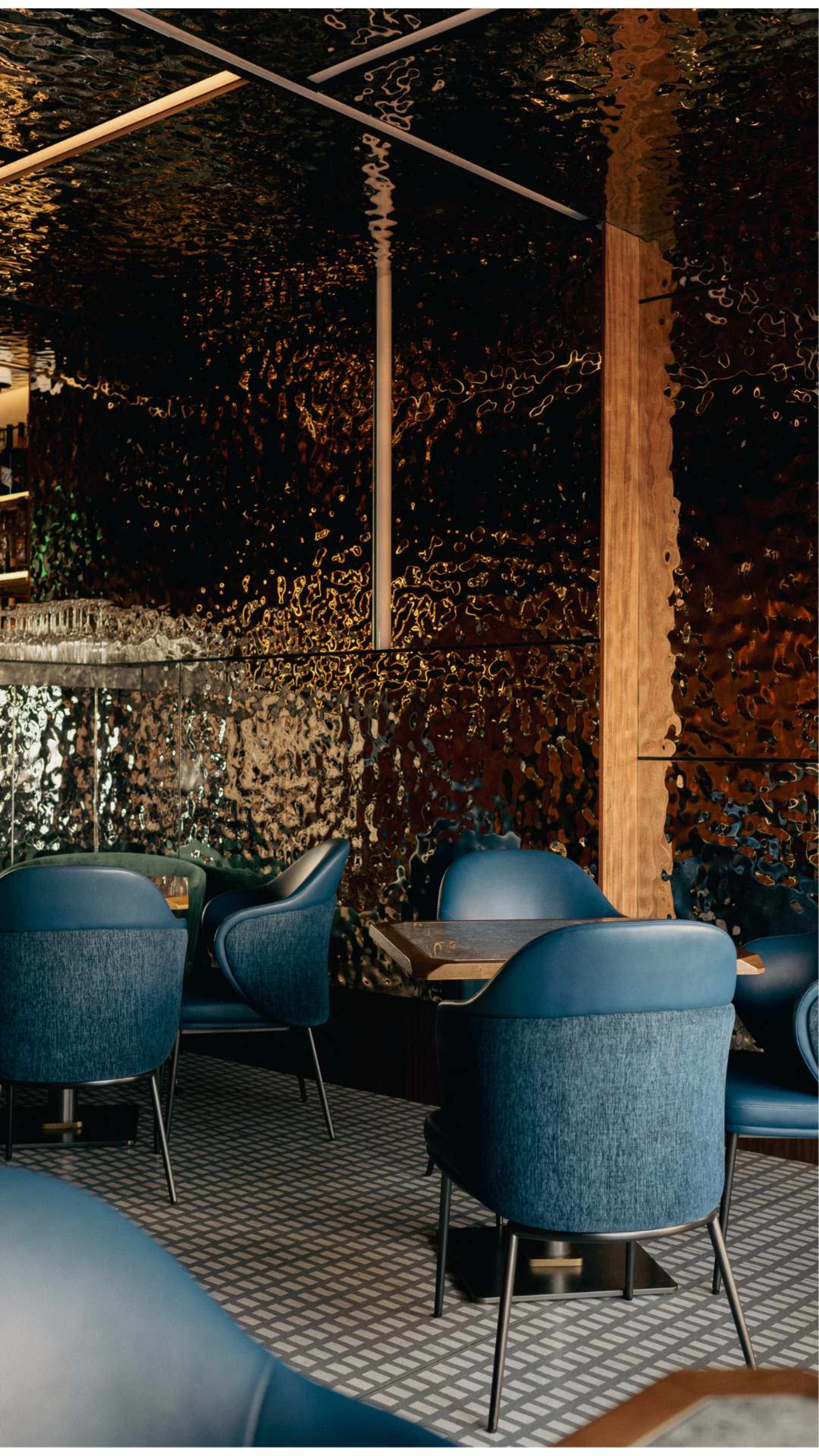 The image shows a modern restaurant interior featuring rounded blue chairs, a geometric patterned floor, and a reflective textured wall.