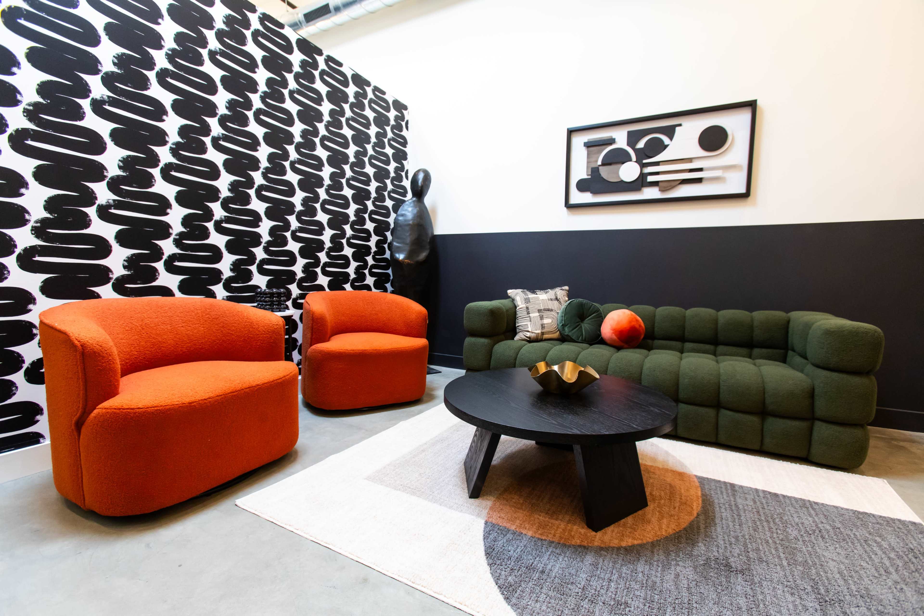 The image features a modern living space with a black and white patterned wall, a green sofa, two orange armchairs, a black coffee table, and a decorative item on the table.