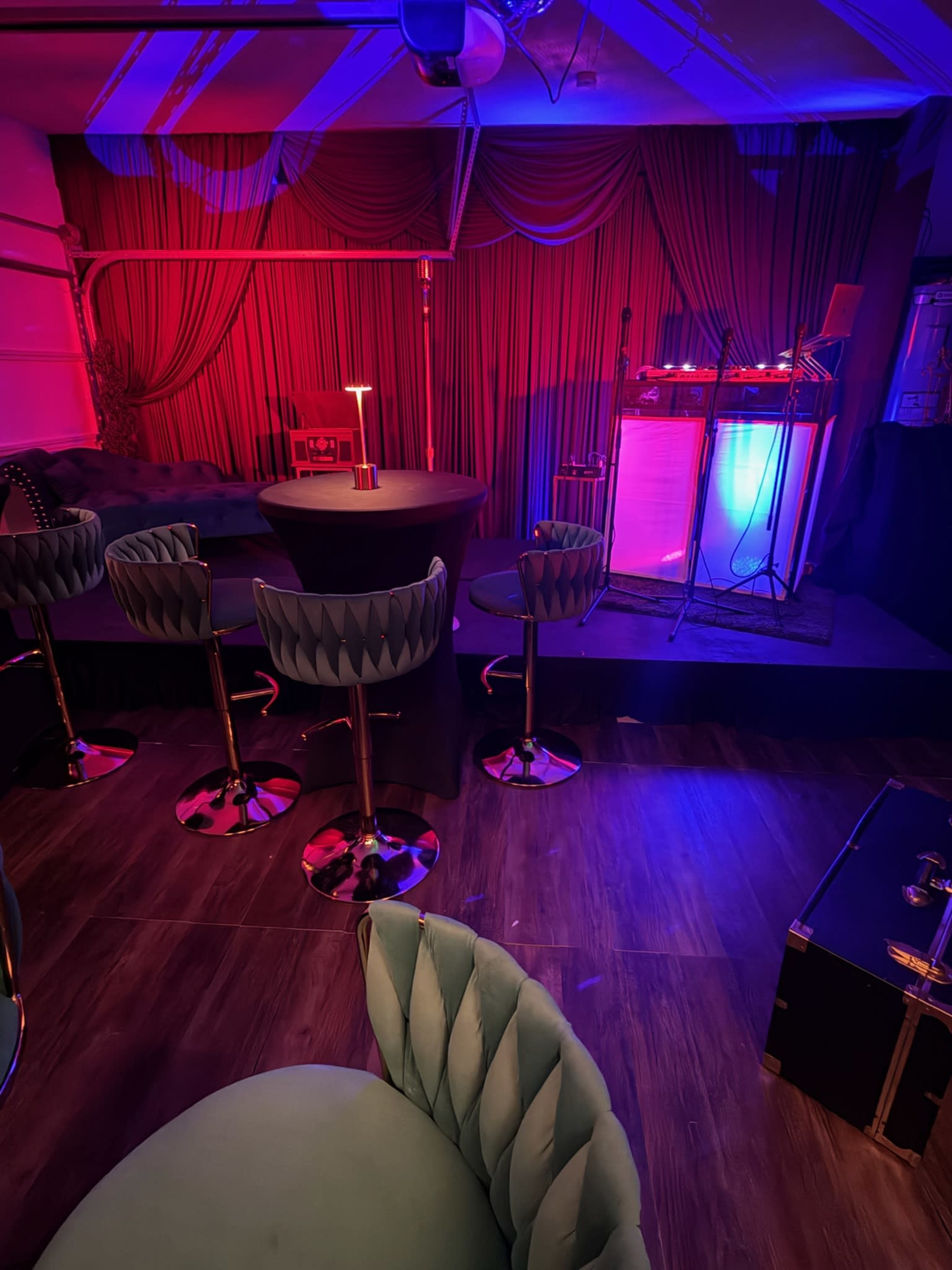 The image shows a dimly lit lounge area with colorful lighting, featuring a bar, several high-backed stools, and a DJ setup in the corner.
