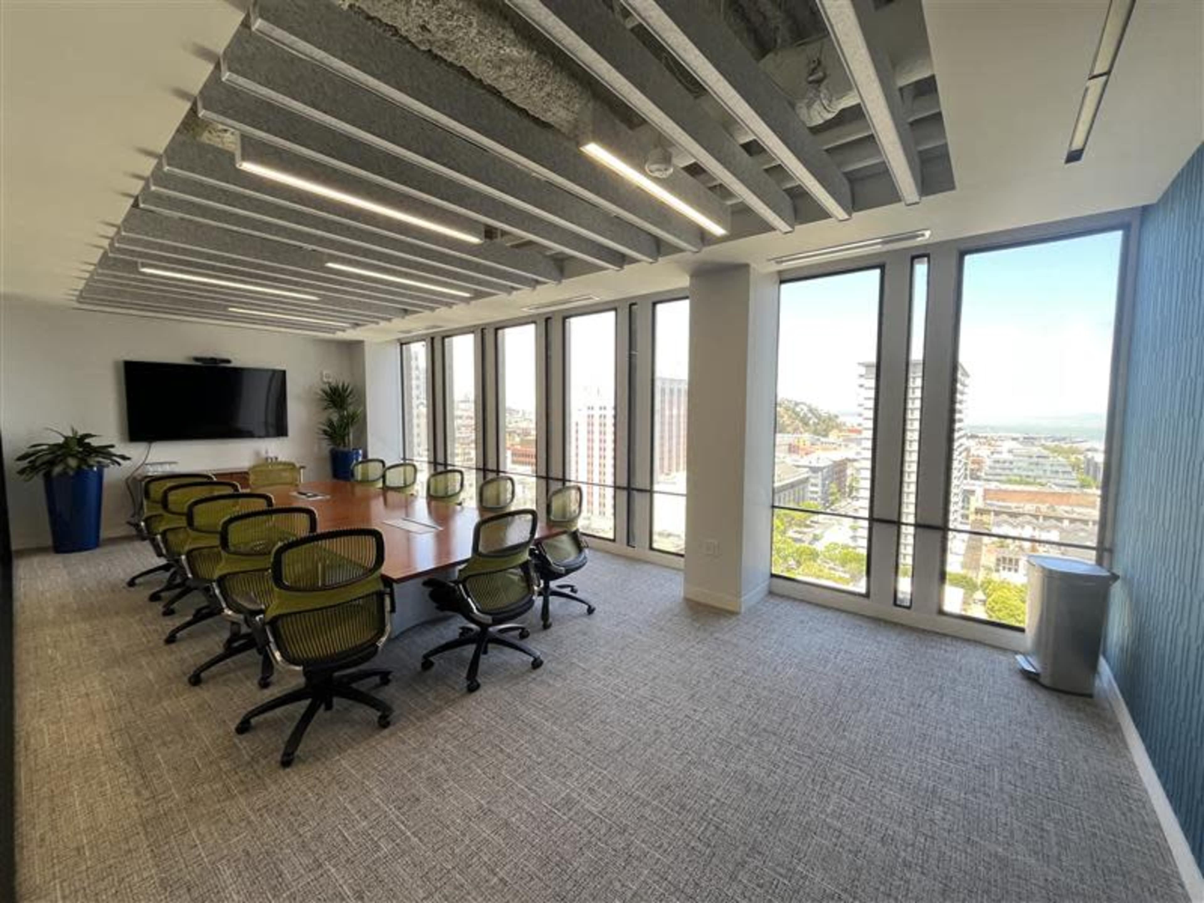 A modern conference room features a long wooden table surrounded by green office chairs, with large windows offering a view of the city skyline.