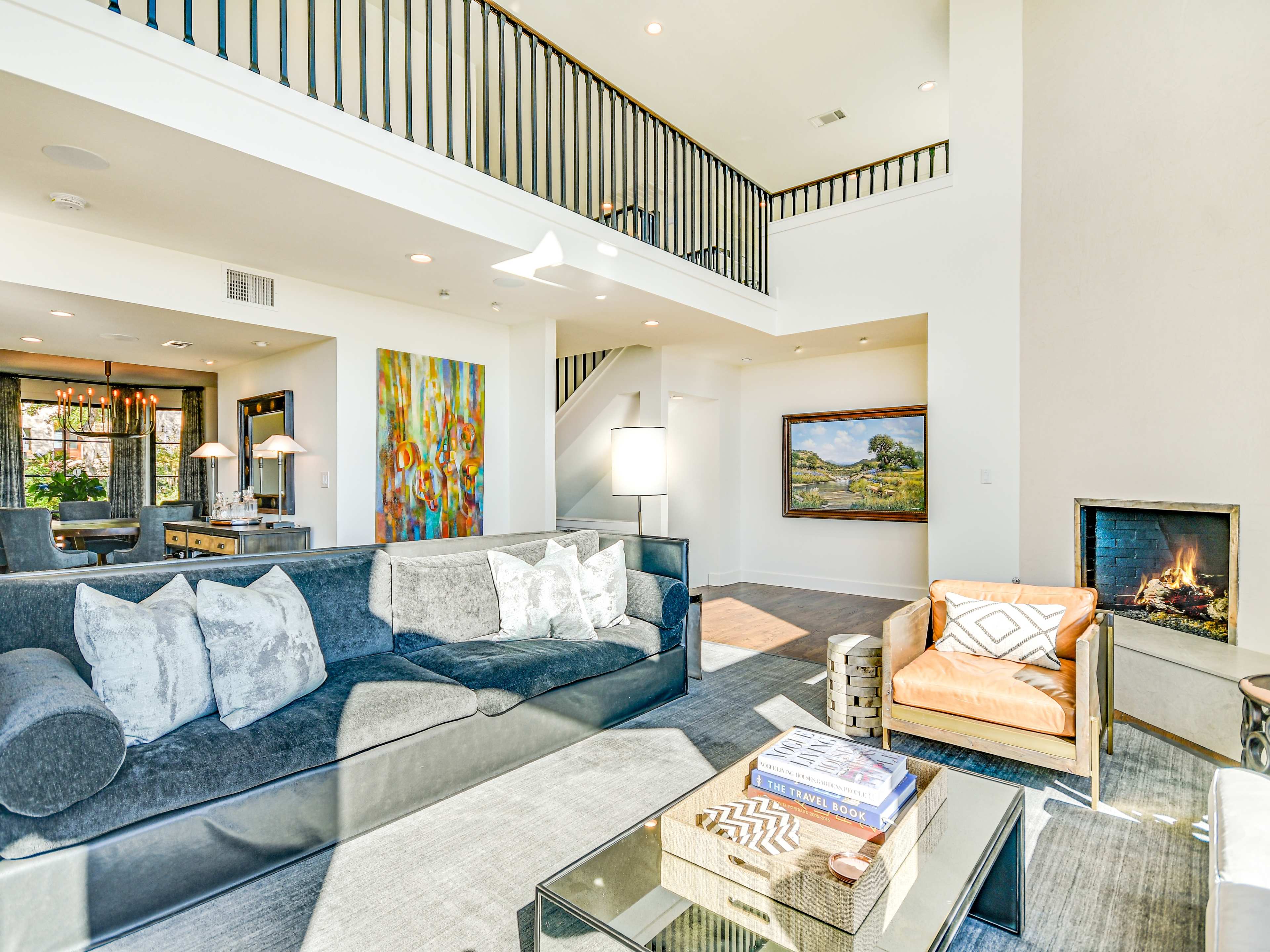 A modern living room featuring a large sectional sofa, an accent chair, a coffee table, and a fireplace, with a dining area and stairs visible in the background.