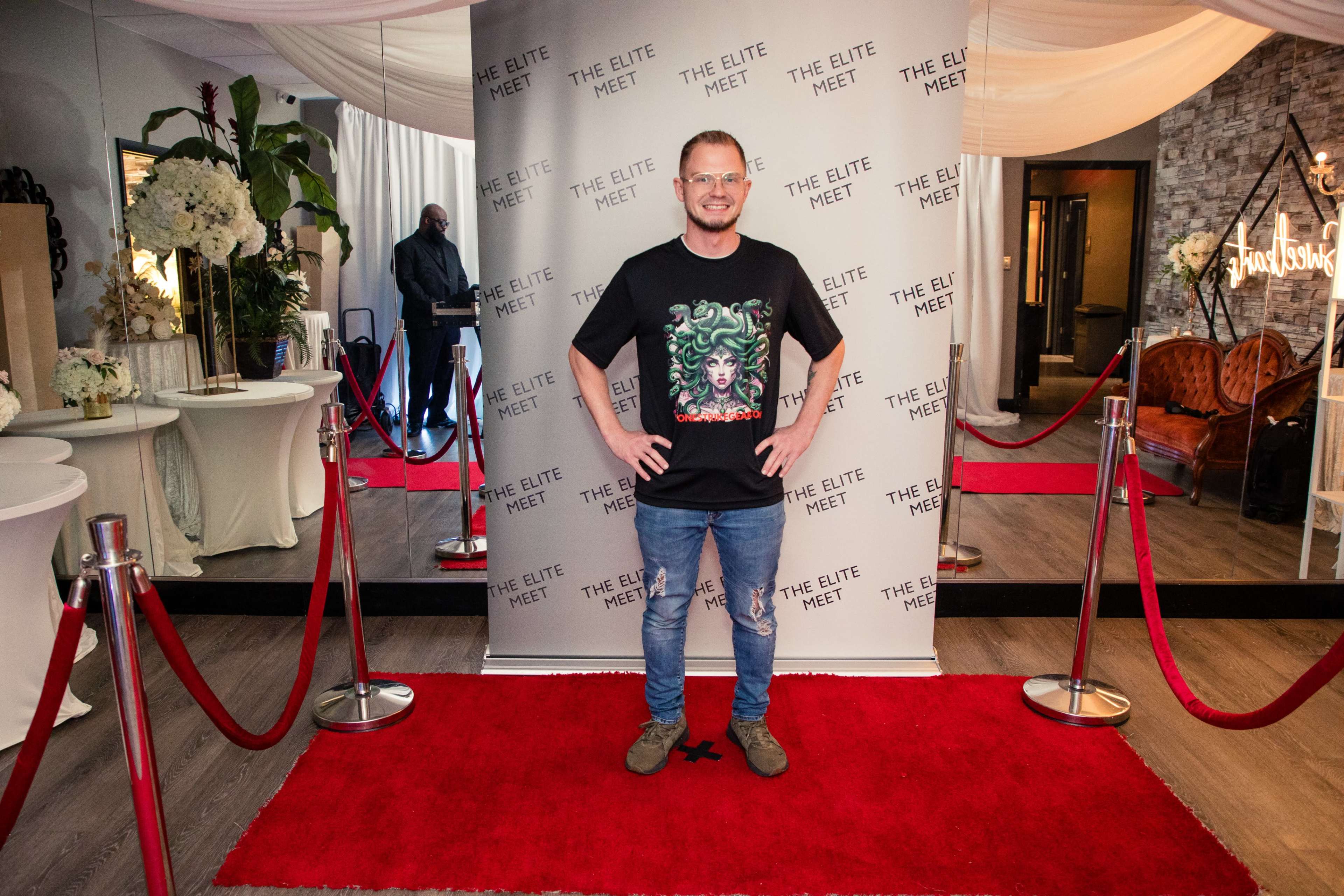 A person stands on a red carpet in front of a branded backdrop at an event venue.