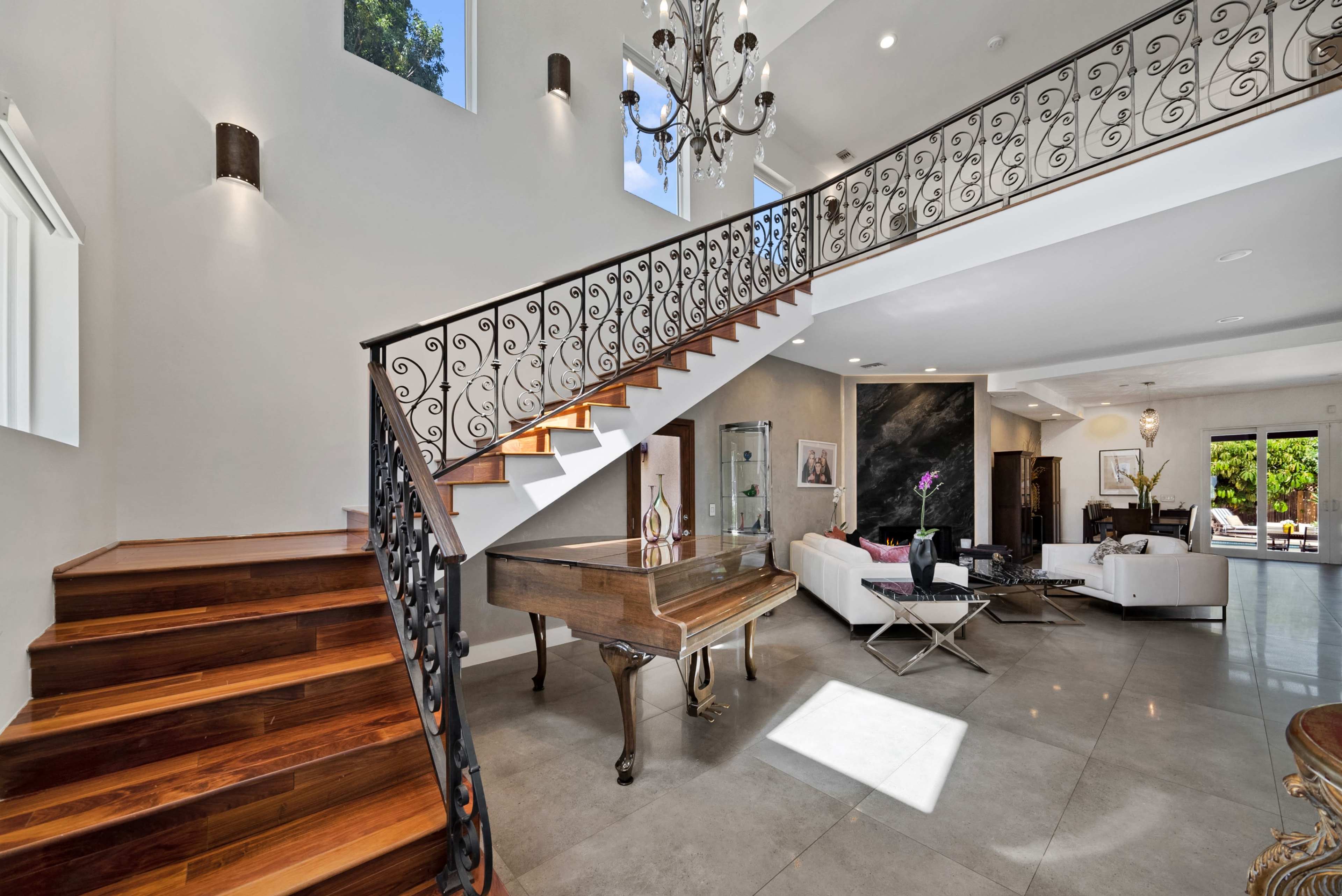 A spacious interior featuring a wooden staircase with an ornate railing, a grand piano, and modern furniture arranged in a light-filled living area.
