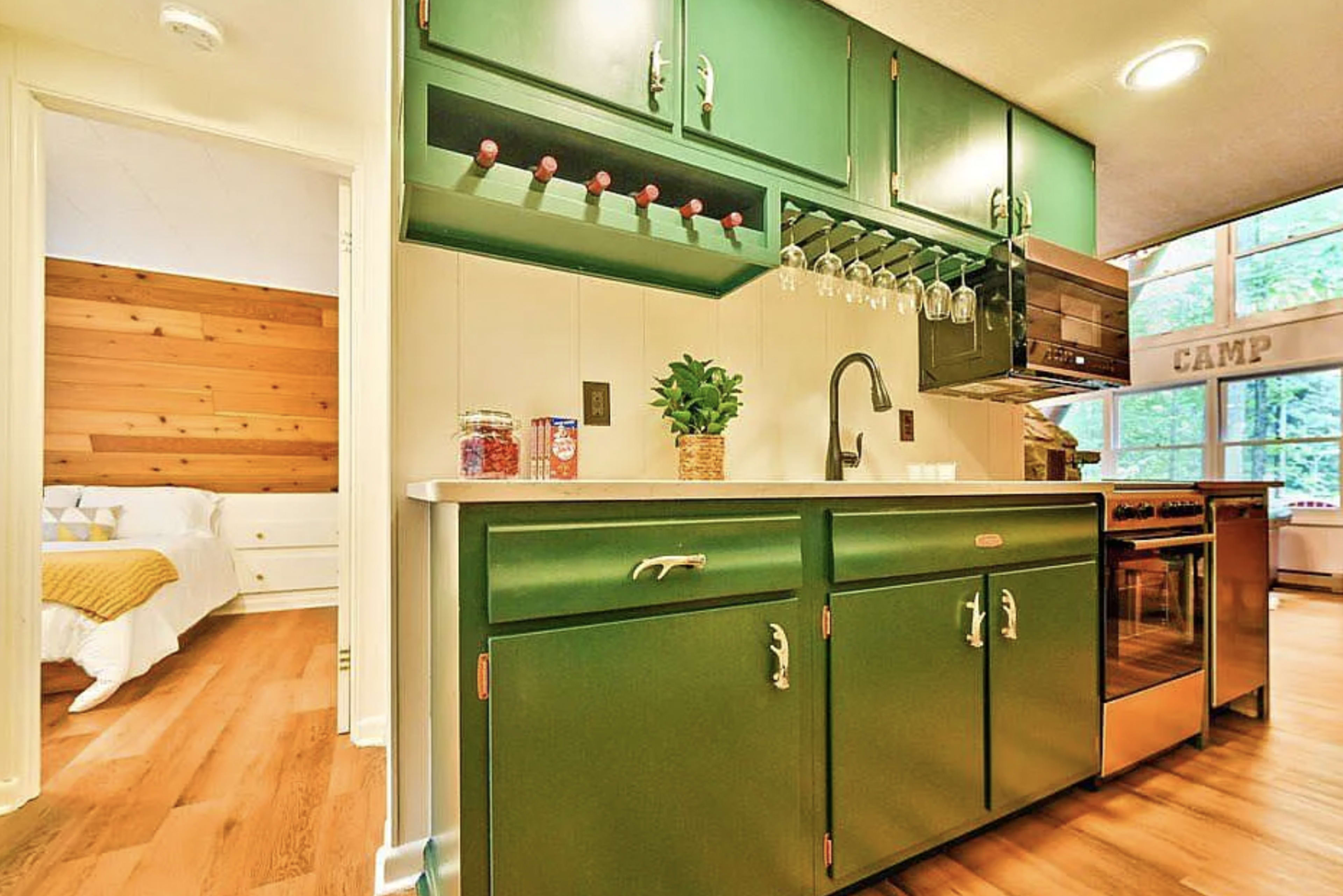 A compact kitchen with green cabinetry, a sink, and stainless steel appliances, adjacent to a bedroom area with wood-paneled walls.