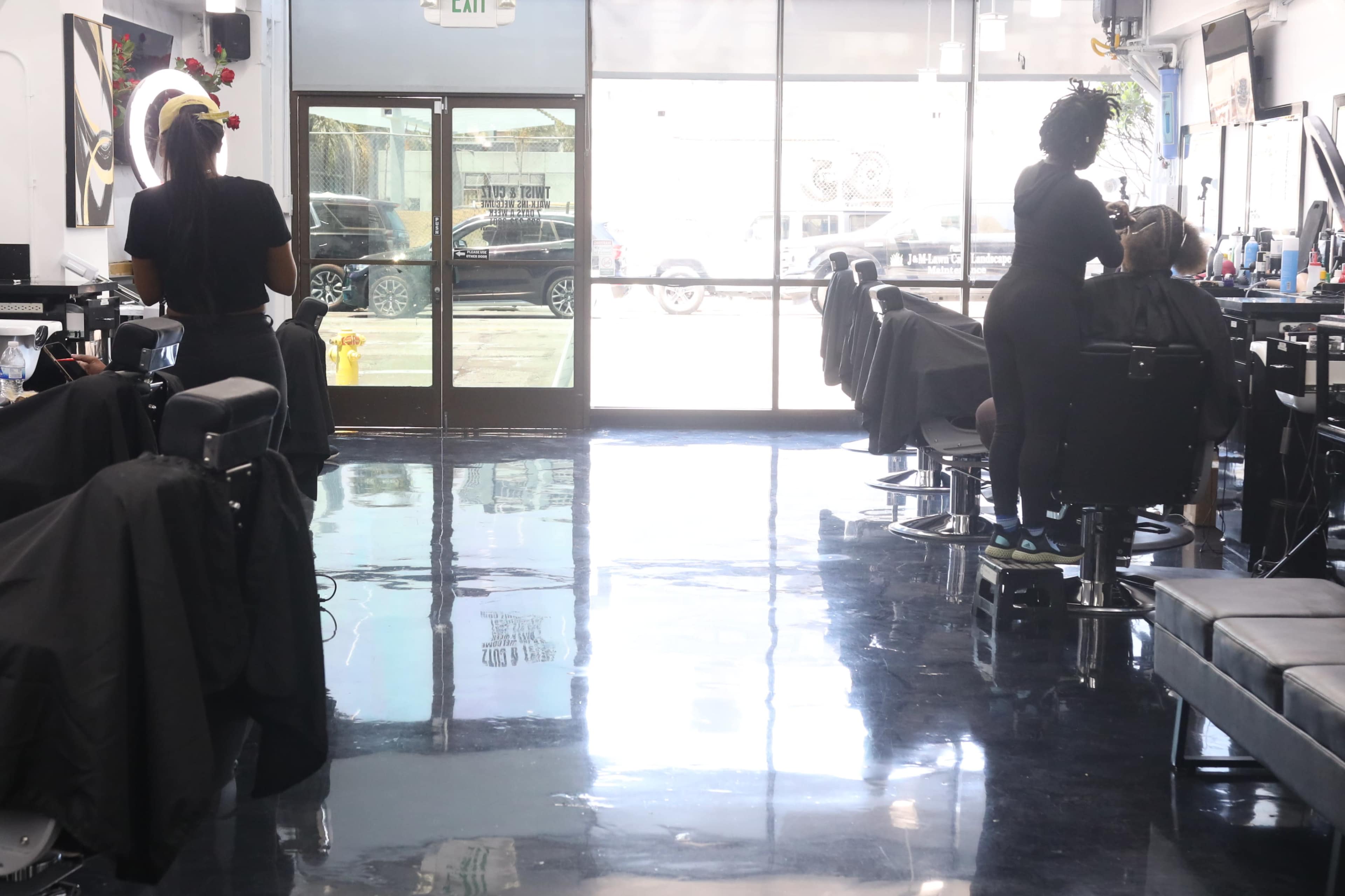 A bright barbershop with two stylists at work, one cutting hair and the other assisting a customer, while sunlight streams through the large front windows.