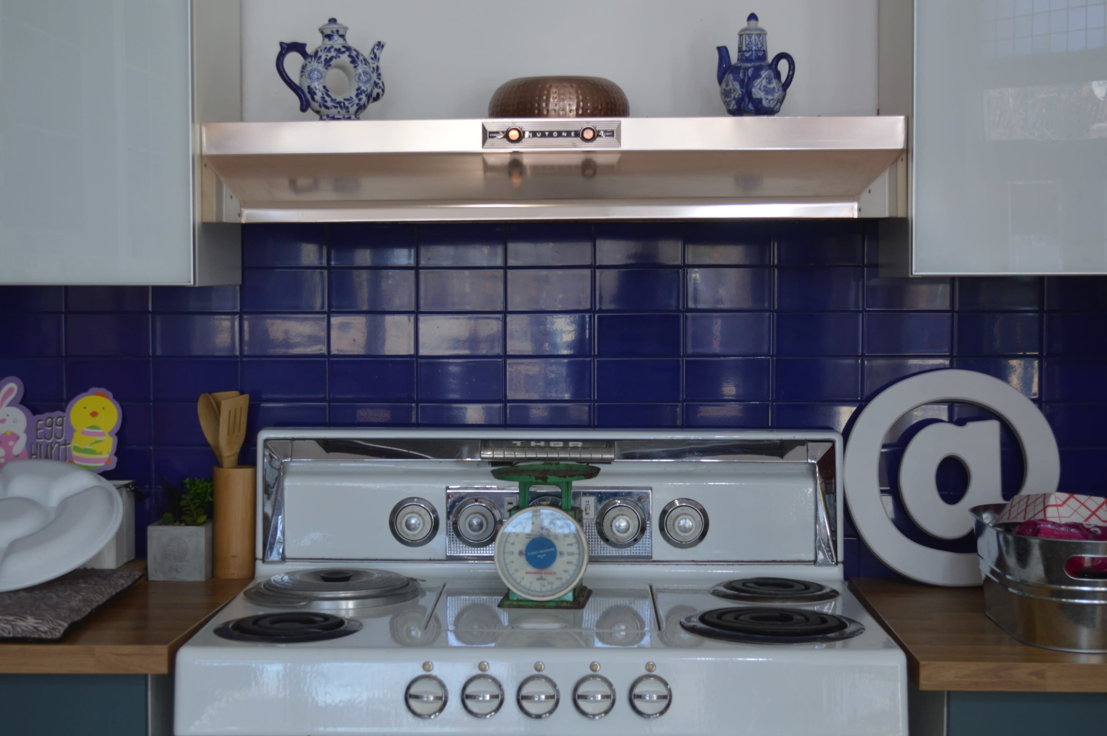 A kitchen stove with a green scale on top and blue tiled walls, adorned with decorative items on the shelf above.