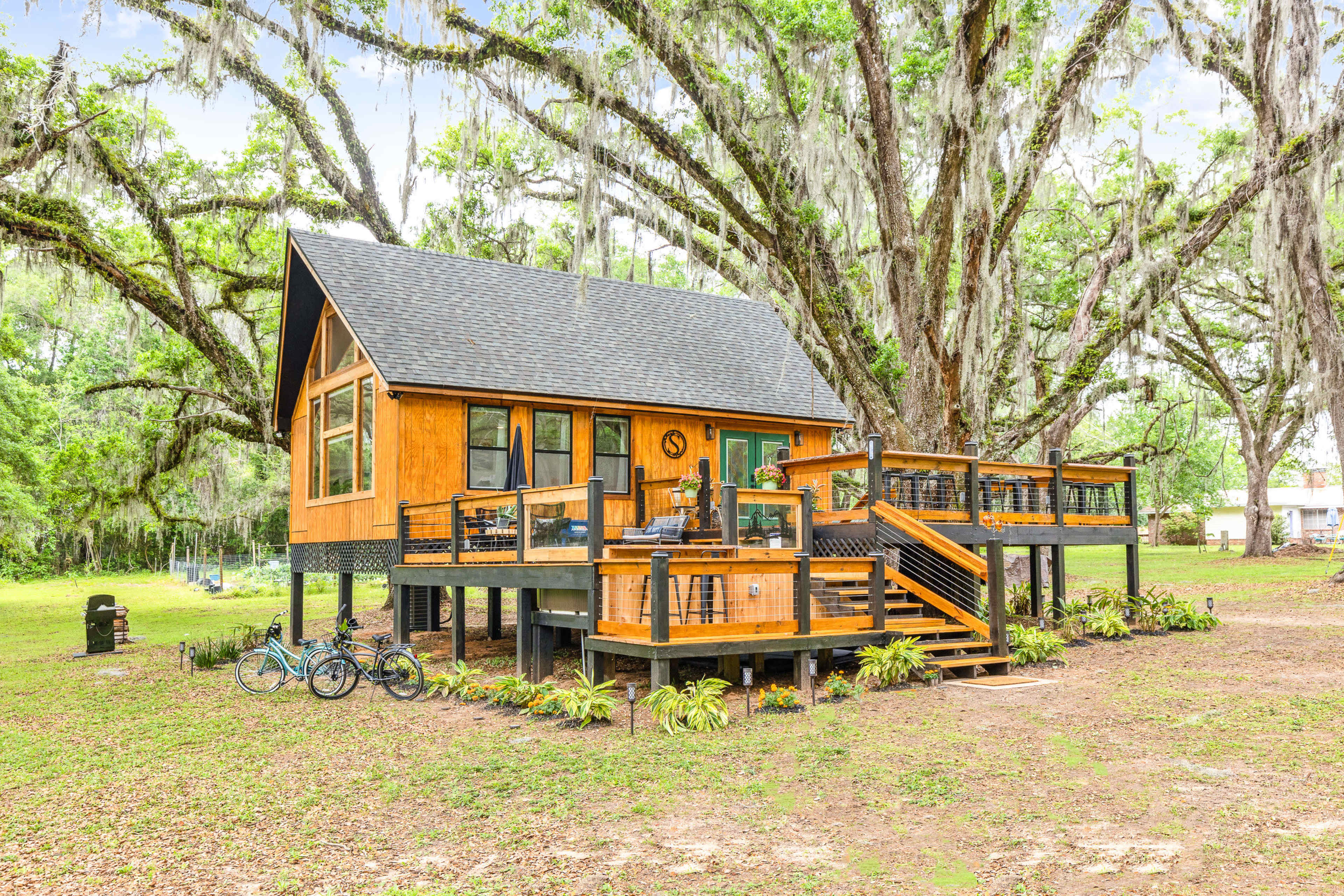 A wooden cabin with a sloped roof is elevated on stilts beneath large, moss-draped trees, featuring a wraparound deck and two bicycles parked nearby.