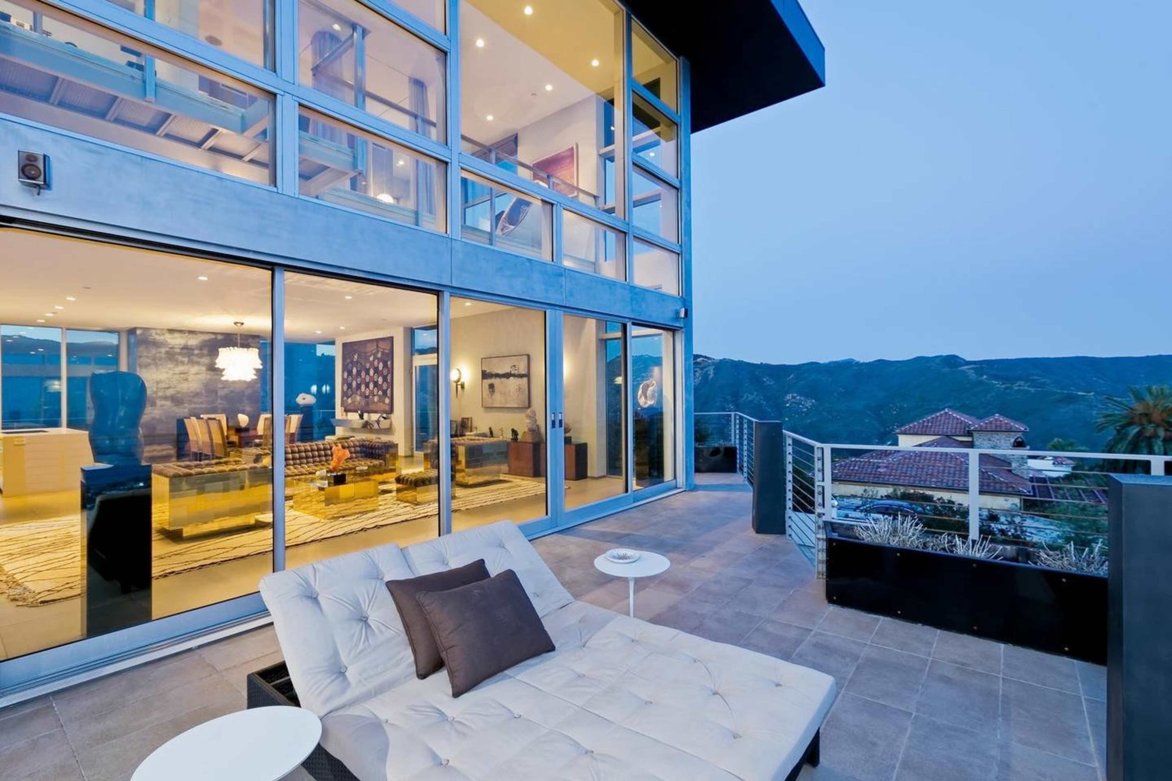A modern house with large glass windows overlooks a mountainous landscape during dusk, featuring a patio with a lounge chair and small table.
