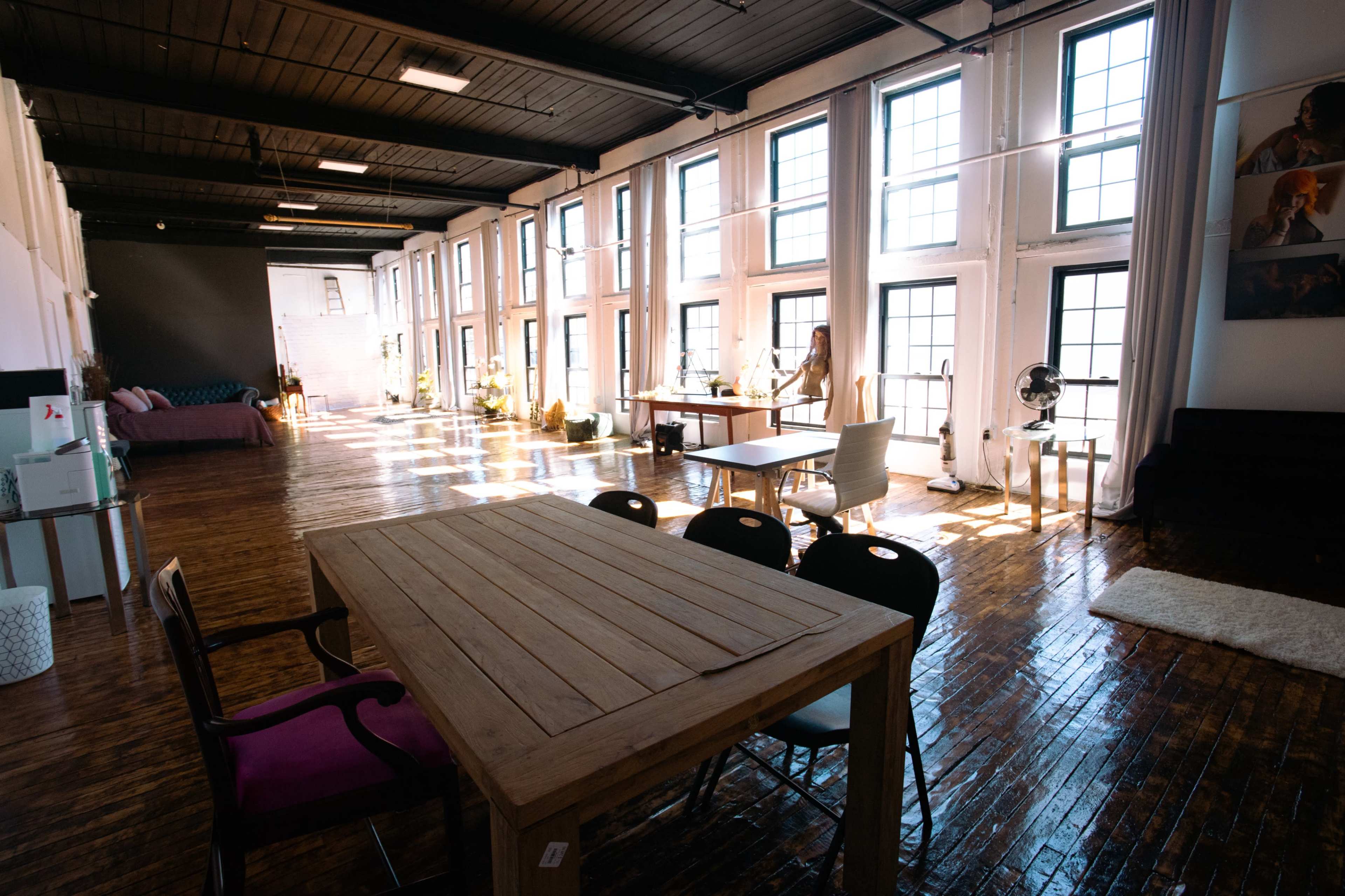 A spacious, well-lit room with large windows, wooden floors, and various furniture items, including a table, chairs, and a sofa.