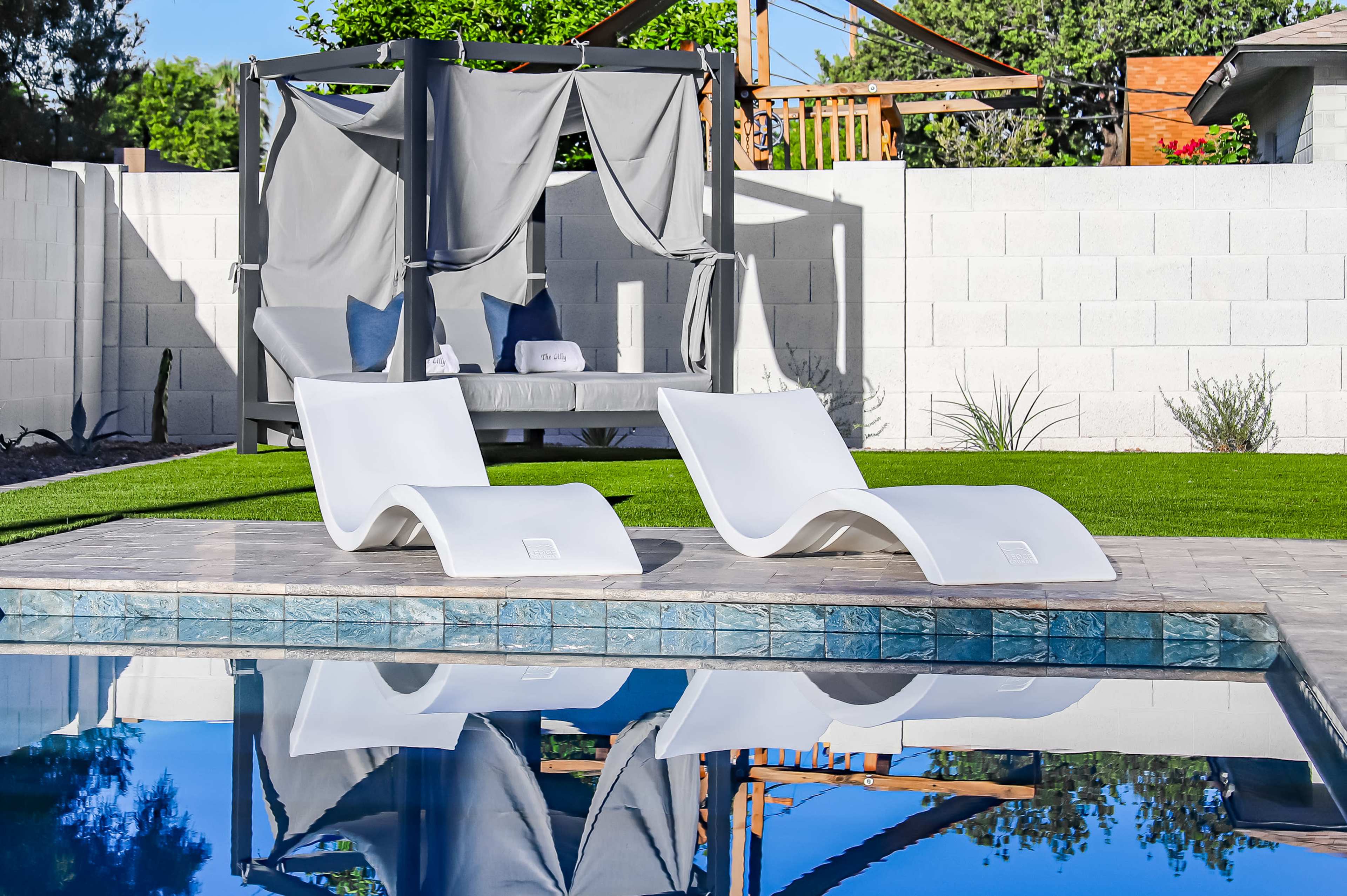 Two white lounge chairs beside a swimming pool, with a canopy-covered seating area in the background and green grass surrounding the setting.