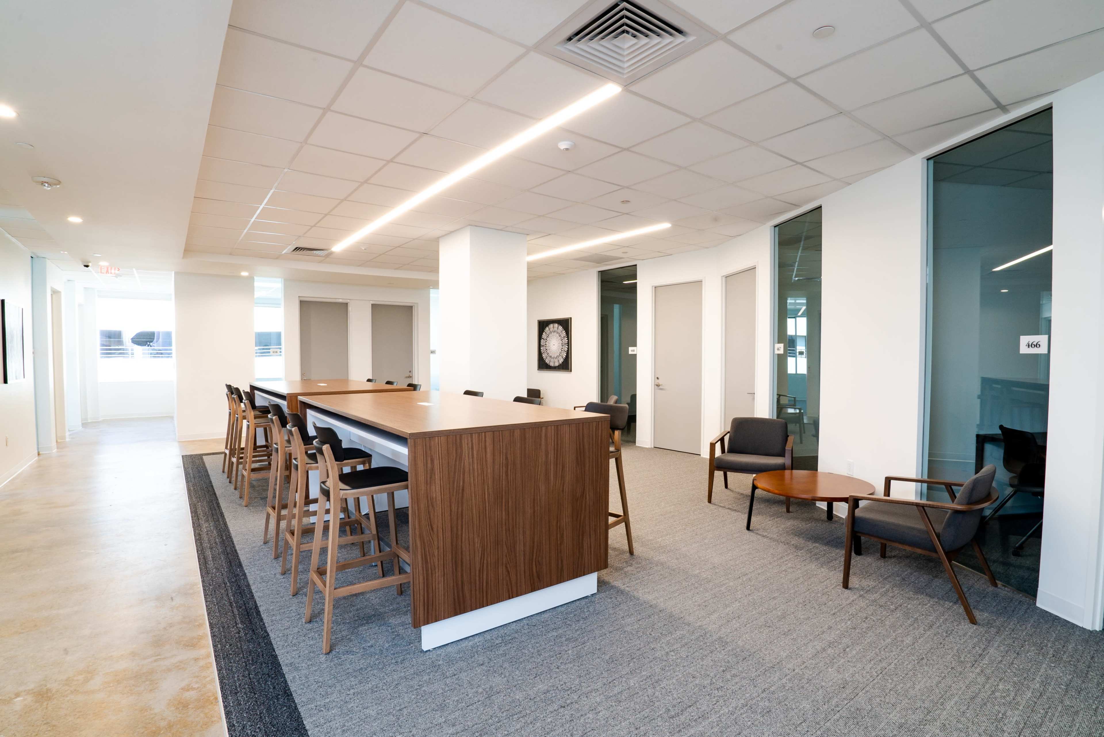 The image shows a modern office space featuring a long wooden table surrounded by high chairs, with additional seating in the corner and large windows providing natural light.