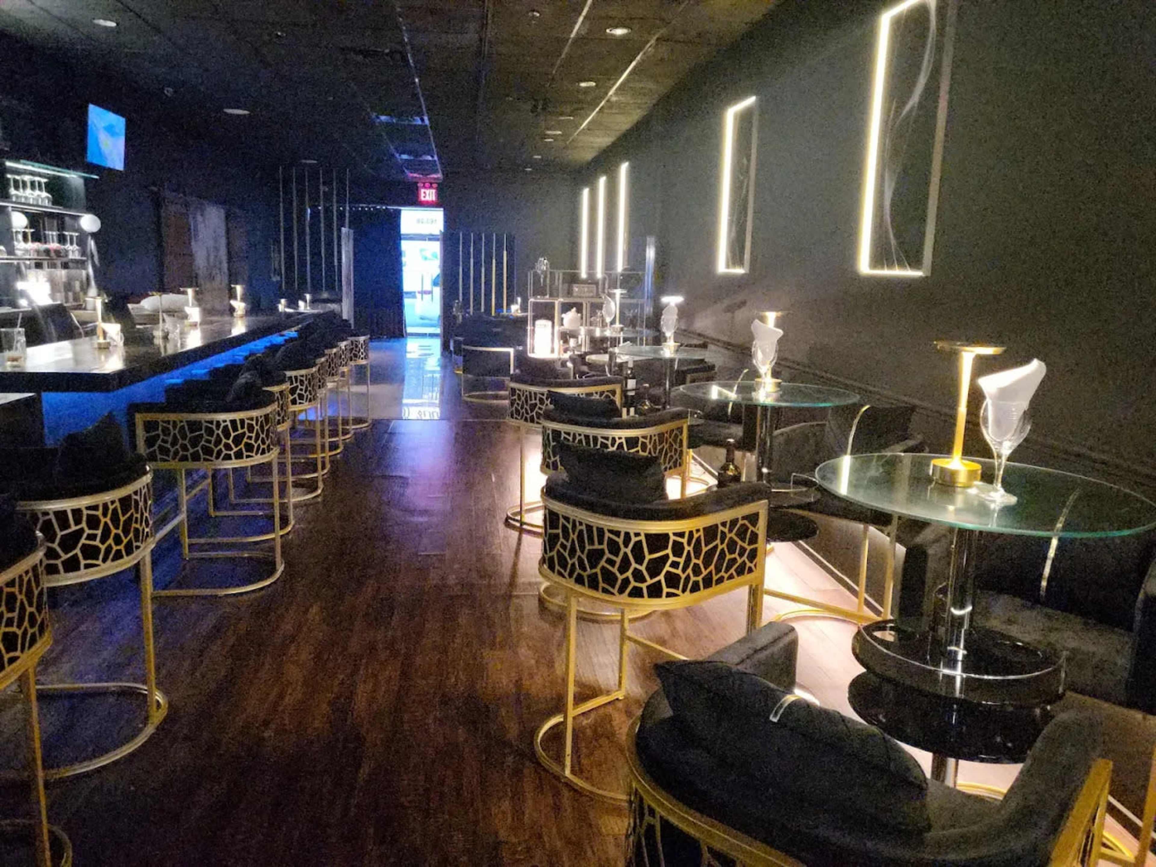A modern lounge features a long bar with stylish seating arrangements, illuminated by soft lighting.