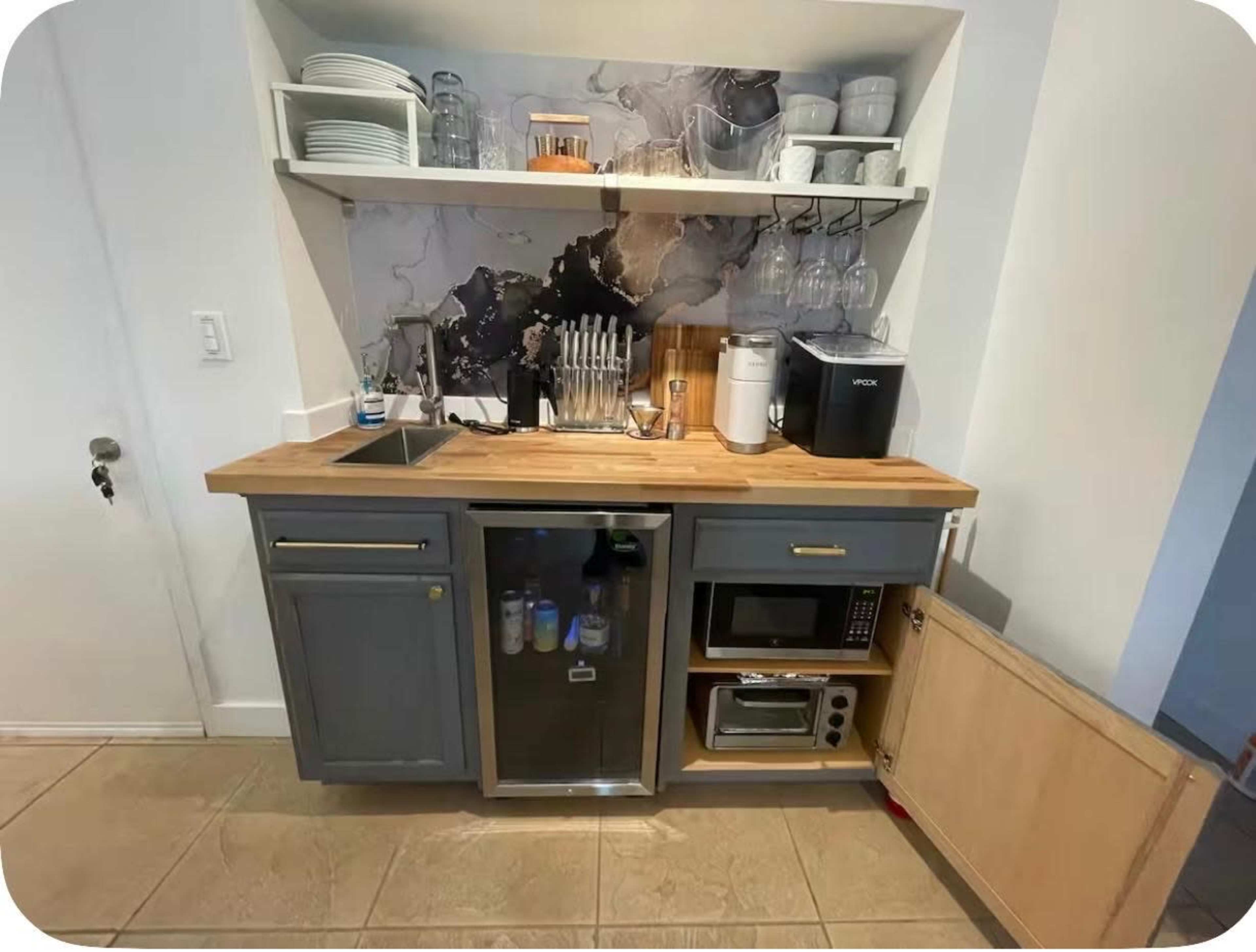 The image shows a compact kitchen area with a wooden countertop, a sink, a beverage fridge, a microwave, and organized glassware on shelves above.