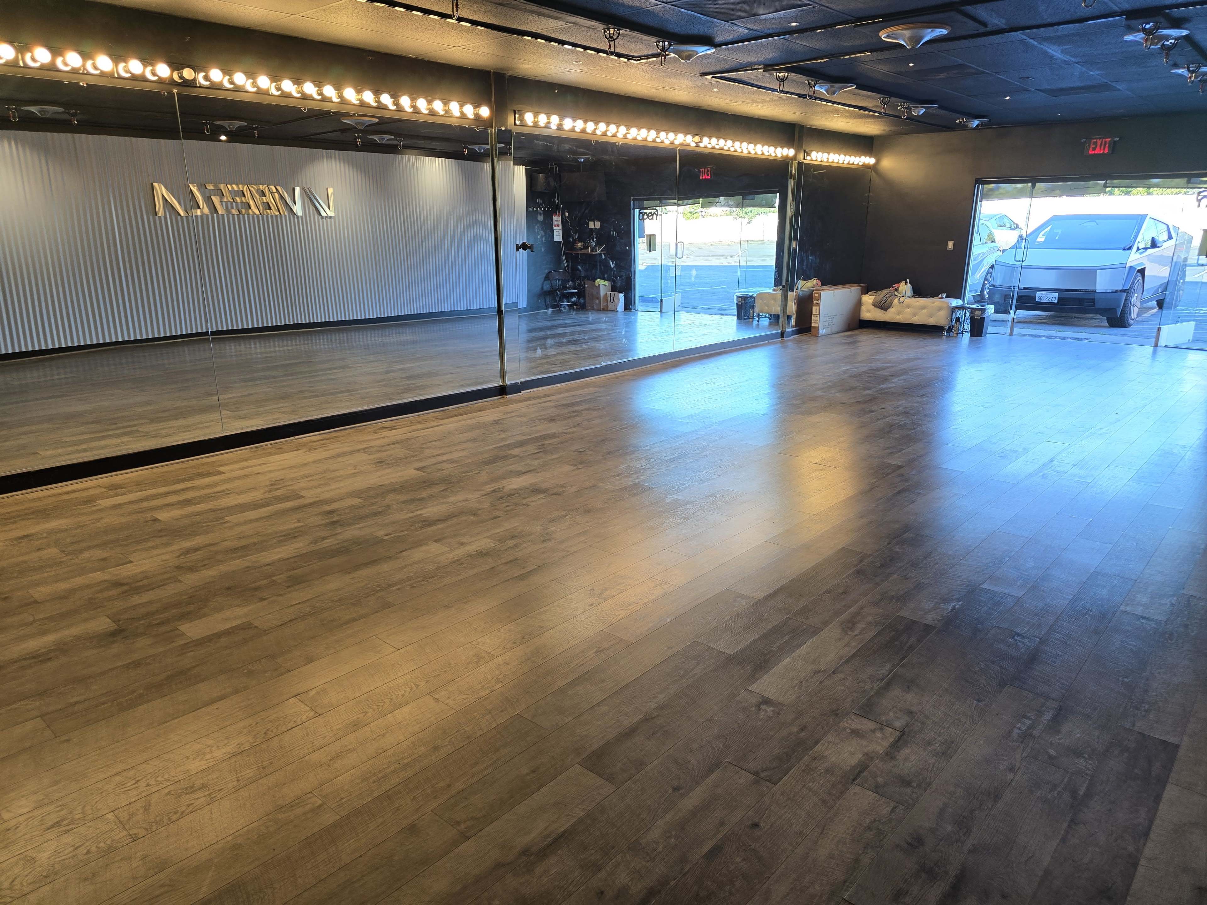 The image shows a spacious studio with wooden floors, a large mirror on one wall, and natural light coming through an entrance.