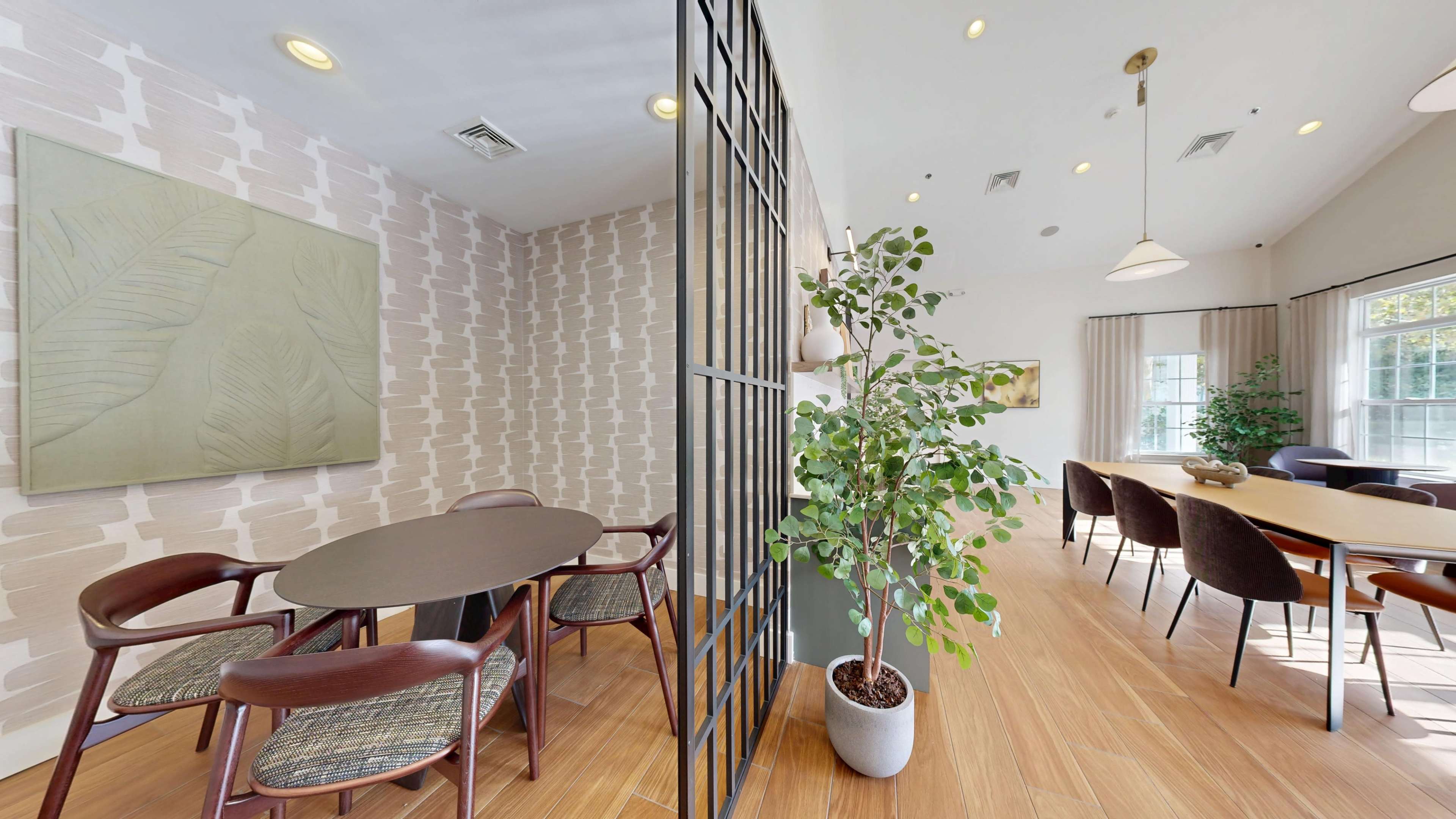 The image shows a modern interior space divided by a black-framed partition, featuring a dining area with a round table and chairs on one side and a larger seating area illuminated by natural light on the other.