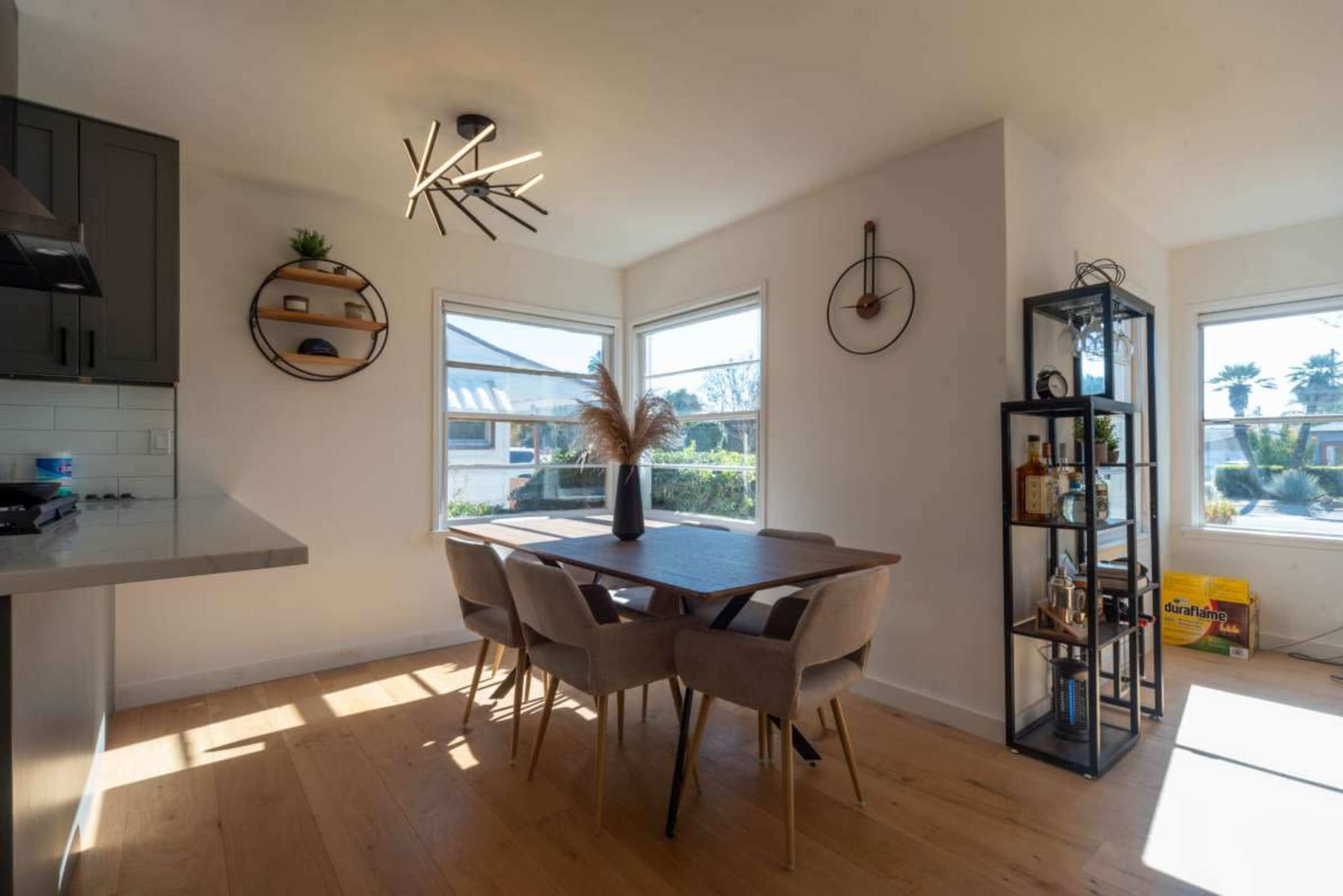 A dining area features a wooden table with beige chairs, ample natural light from large windows, and modern decor elements including a light fixture and wall clock.